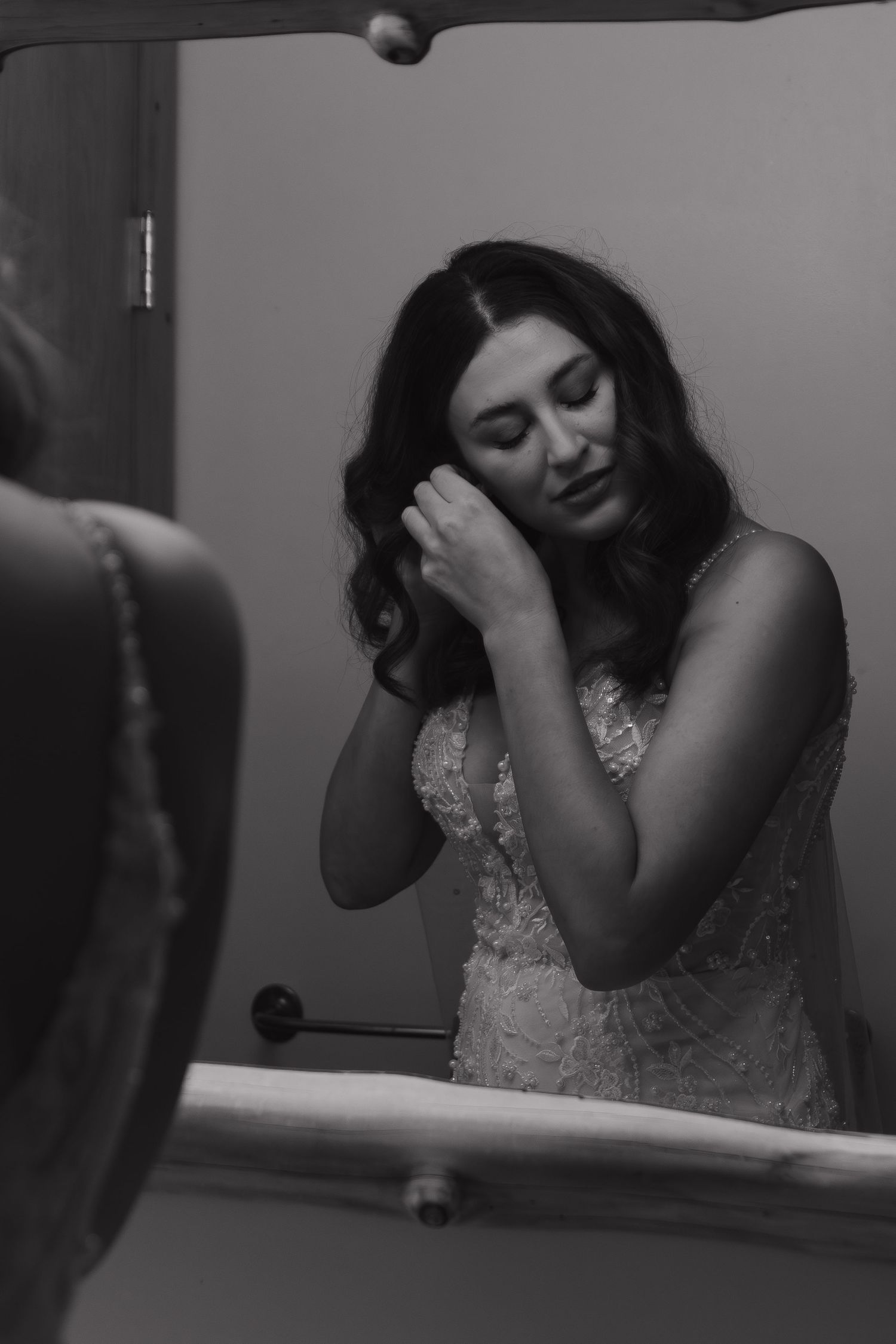 A black and white portrait of a bride in a lace dress looking down while getting ready in front of a mirror.