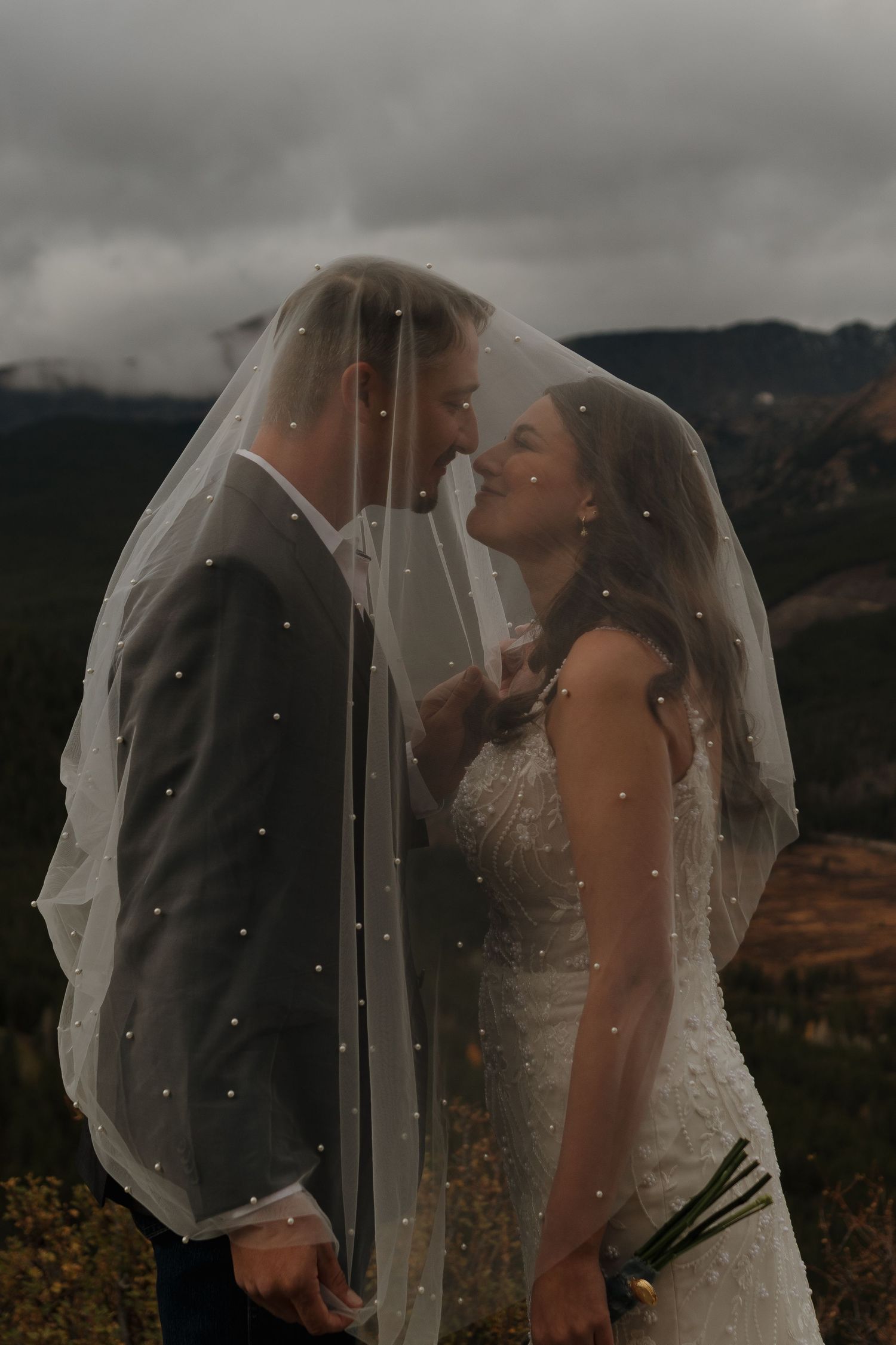 Newlyweds share a tender kiss beneath a sparkling veil against a moody mountain backdrop.