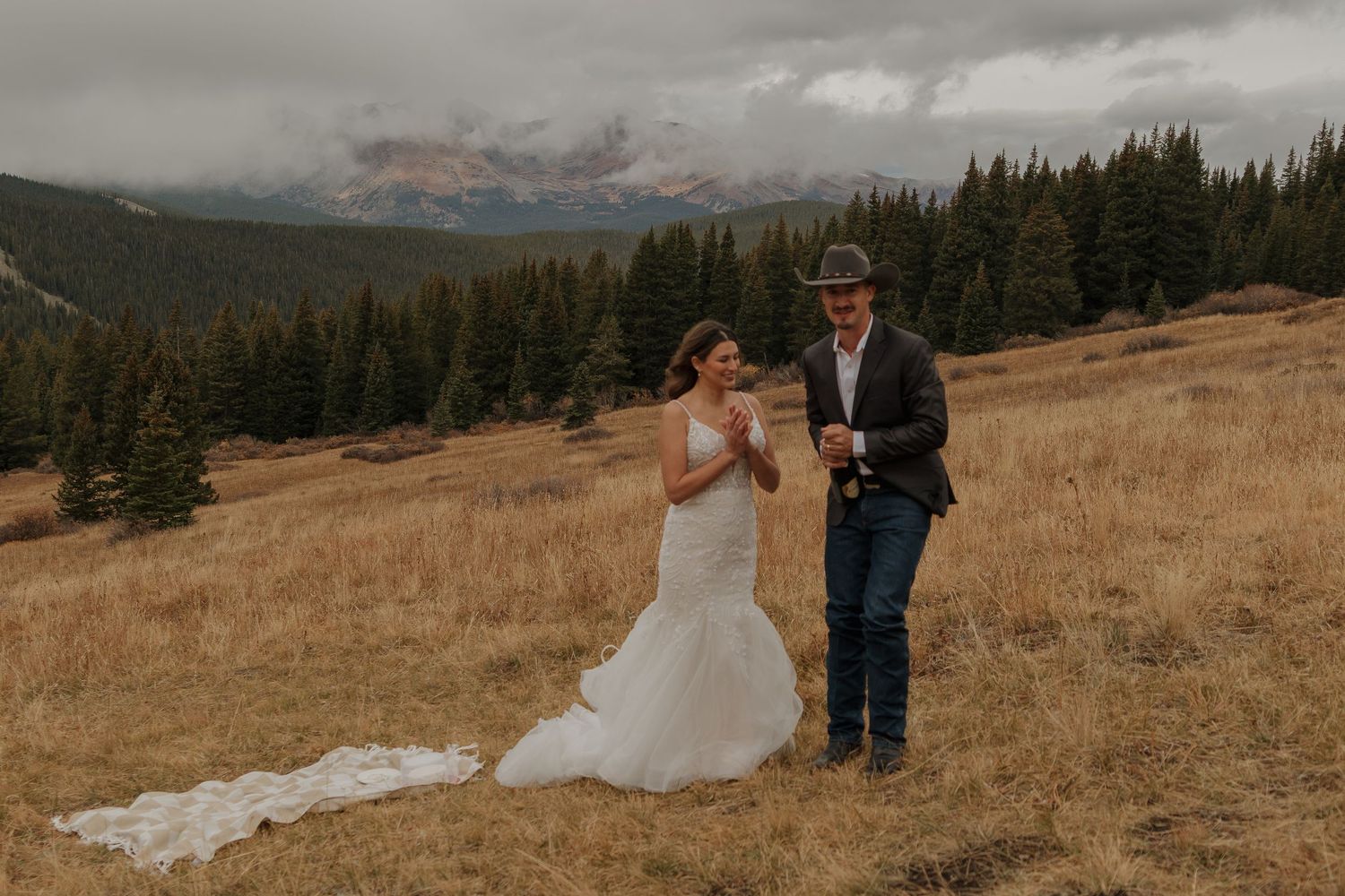 A couple celebrates their wedding on a mountain meadow with dramatic cloudy skies and pine forests.