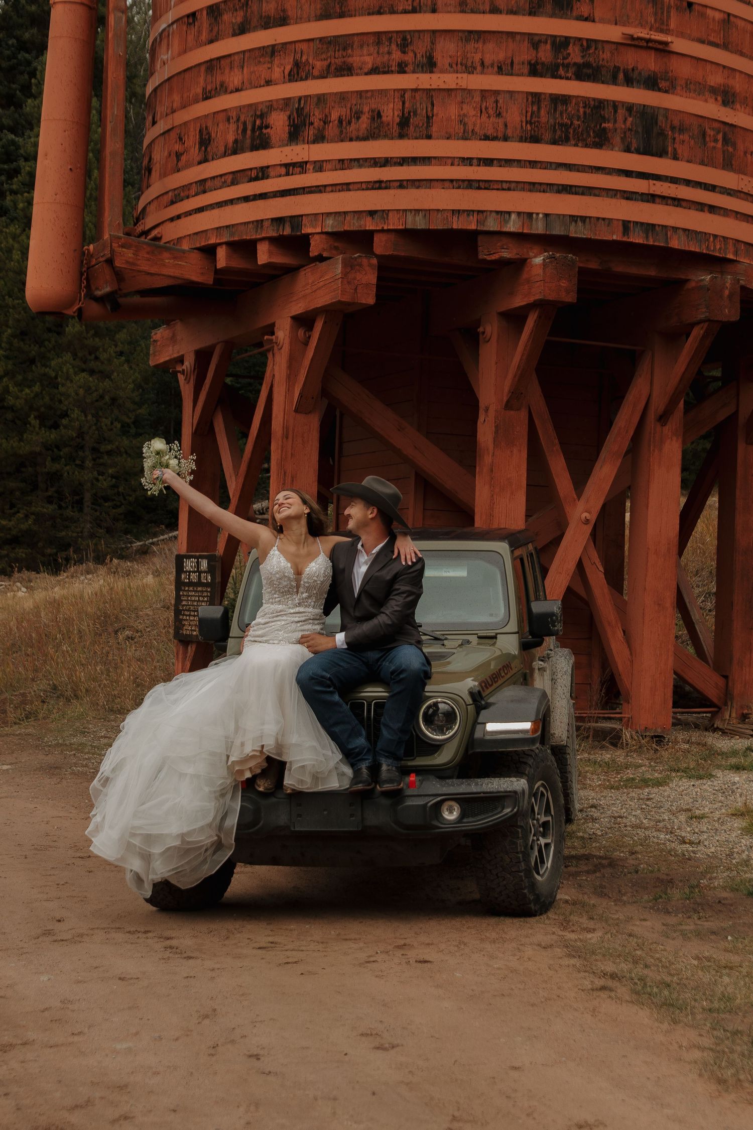 A rustic wedding photo featuring a couple posed on a Jeep near a red wooden water tower.
