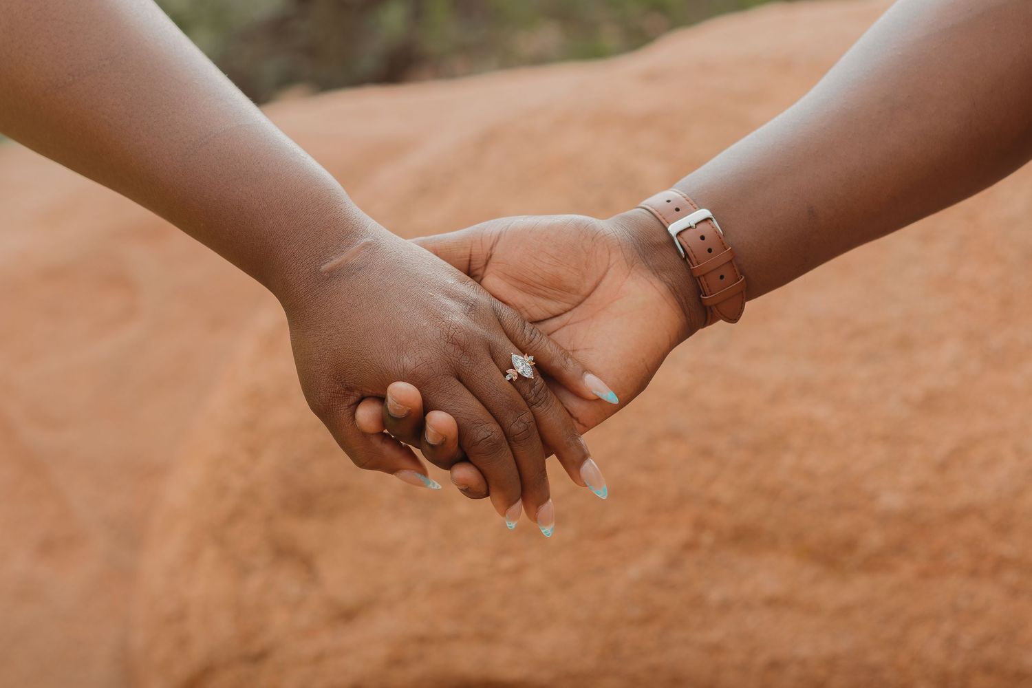 Two hands wearing rings hold each other against a warm-toned sandy or desert background.