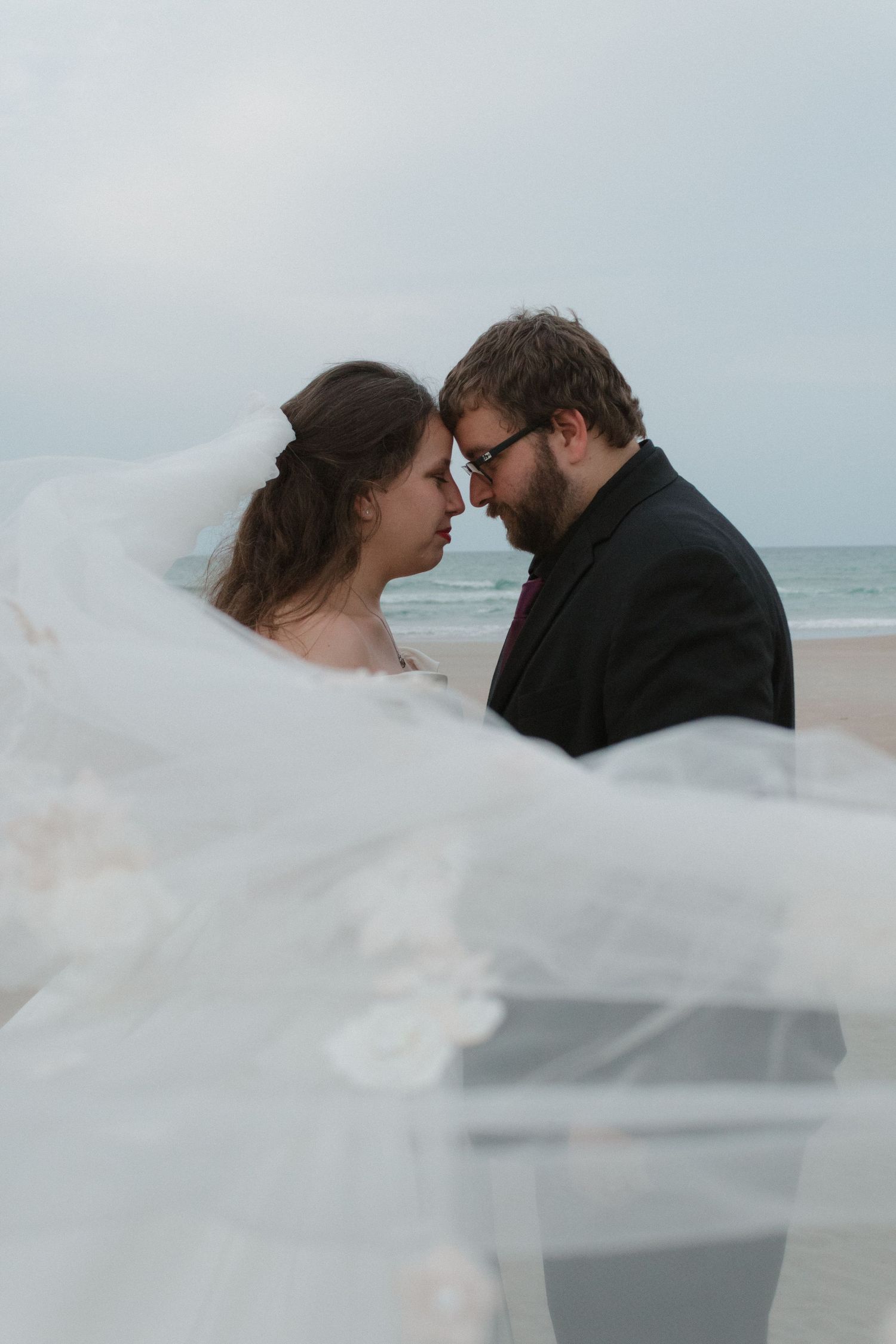 A romantic beach wedding portrait with a flowing white veil in the foreground and ocean waves behind the couple.