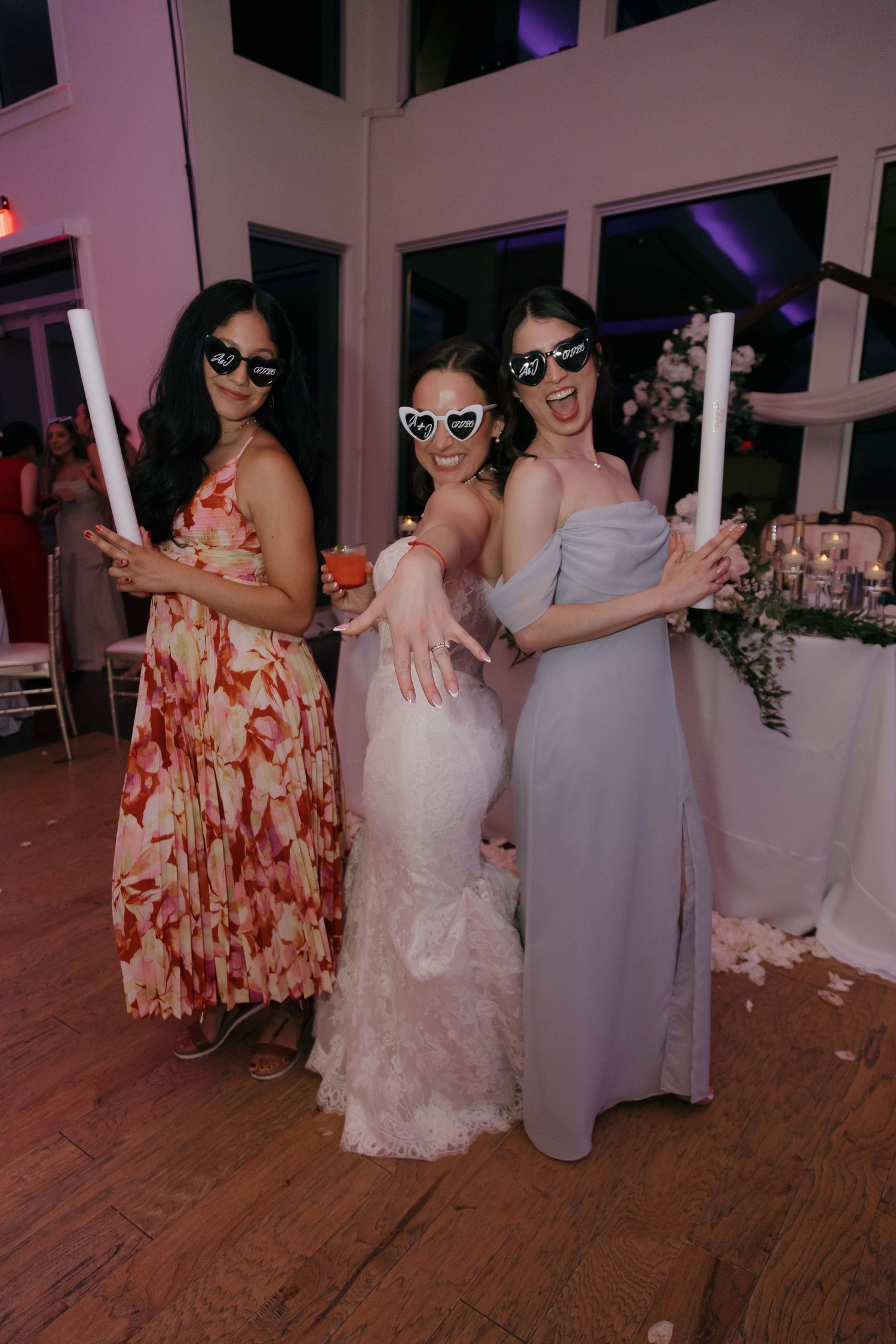 Bride and two guests wearing heart-shaped sunglasses posing with glow sticks at a wedding reception.