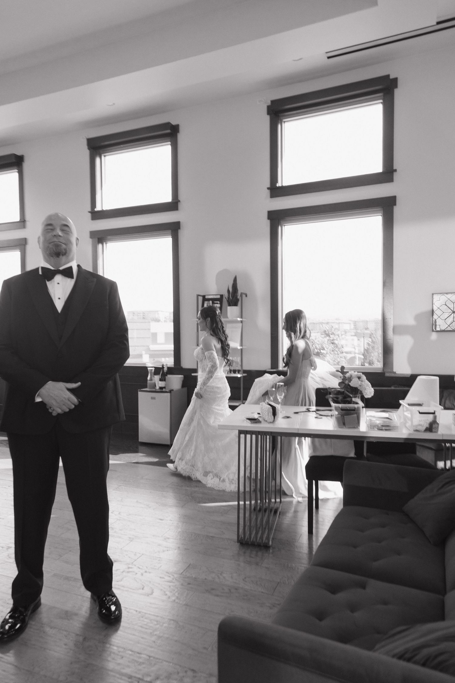 Black and white photo of groom standing while bride prepares with attendant in bright loft space.