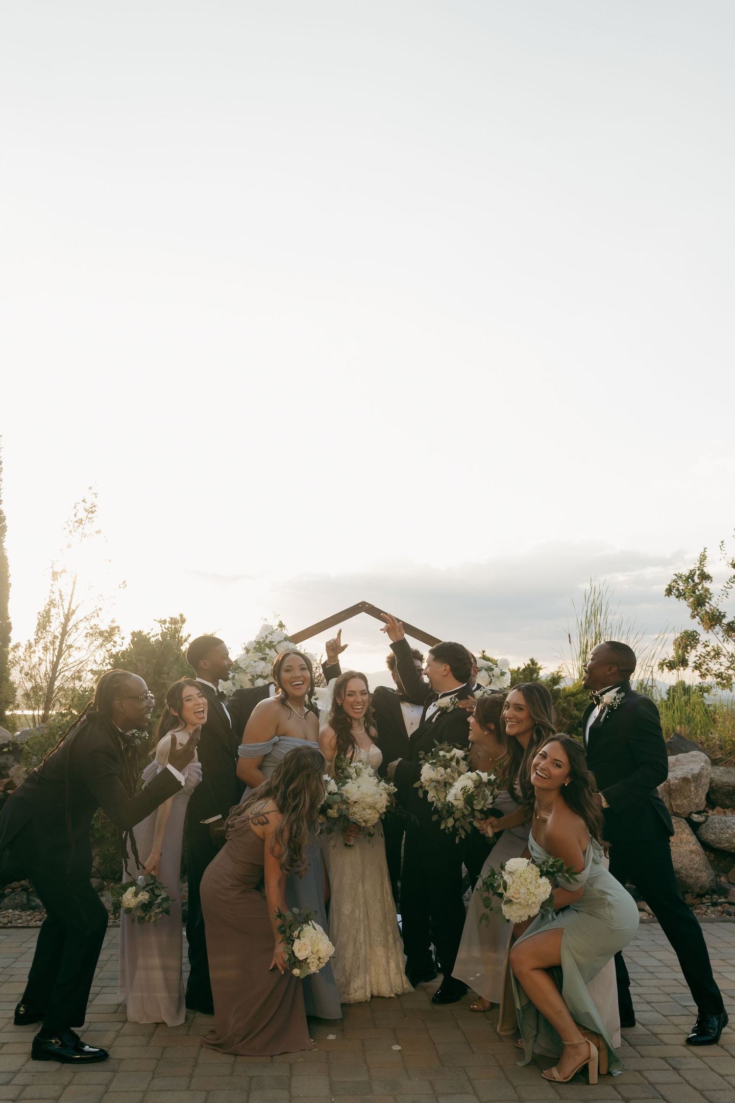 Wedding party celebrates with bride and groom under arch at golden hour outdoor ceremony venue.