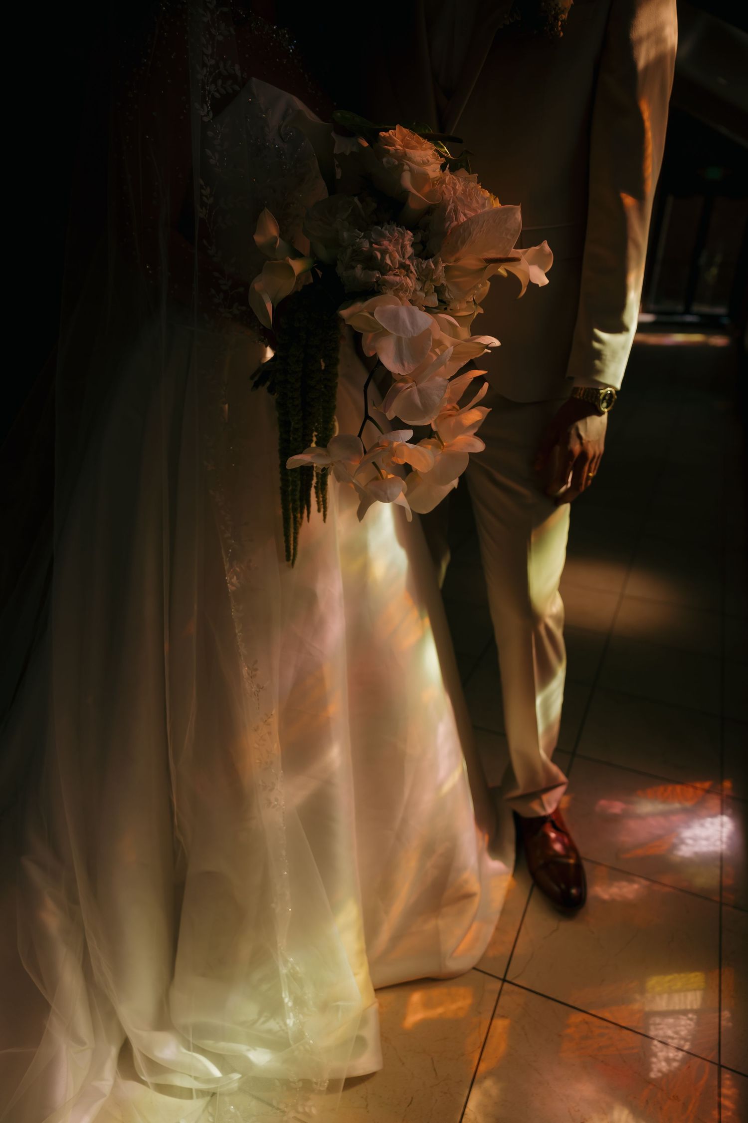 Bride holding pink floral bouquet beside groom in tan suit with stained glass light reflections.