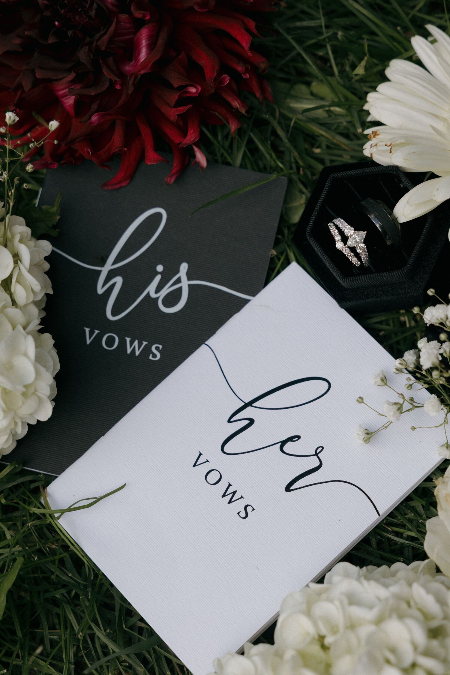 His and her vows books with wedding rings in a velvet box surrounded by white and red flowers.