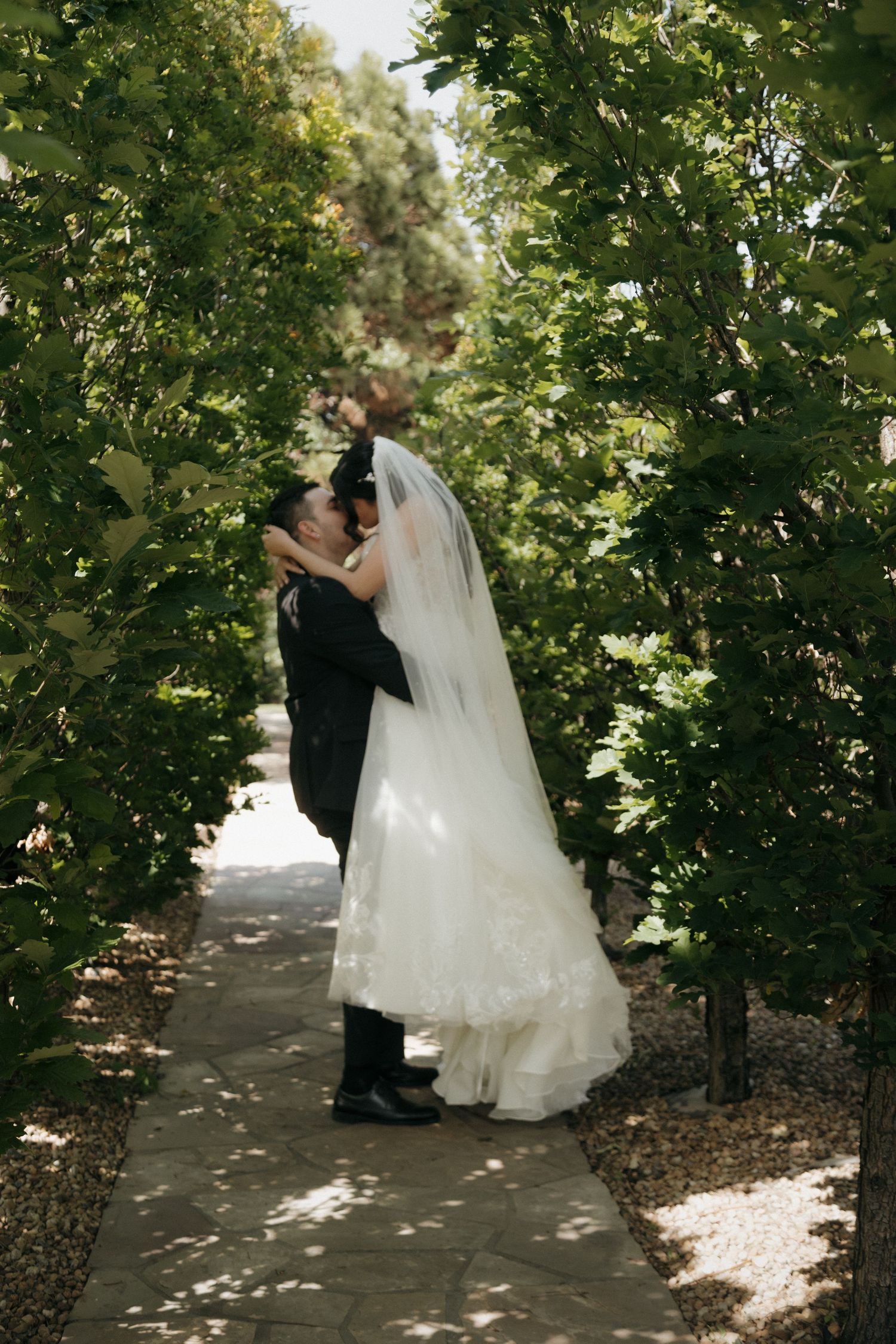 Groom lifts bride in white lace gown for a kiss on a garden pathway lined with green trees.