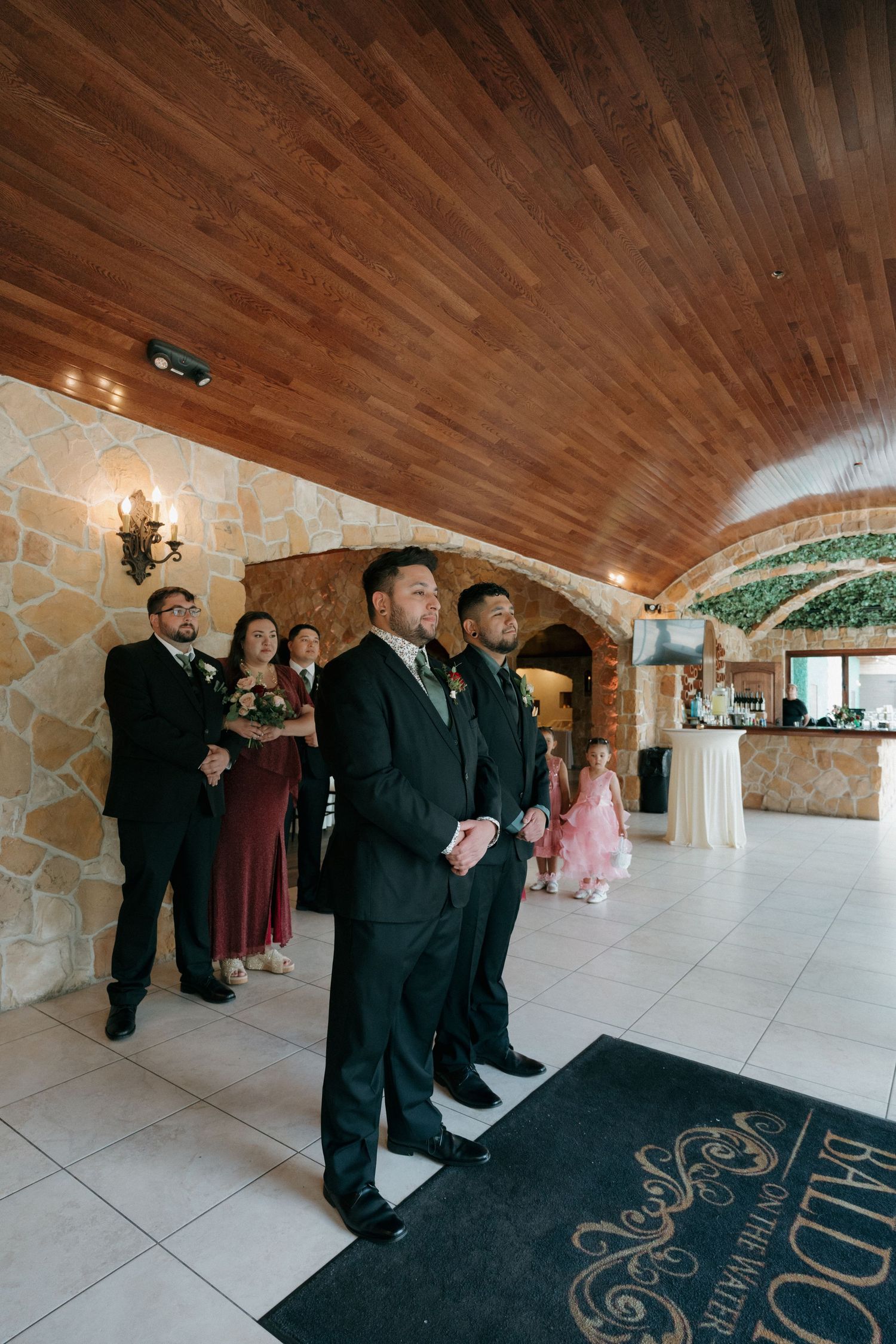 Groom and groomsmen wait inside a stone arch venue as bridesmaids in burgundy dresses stand nearby.