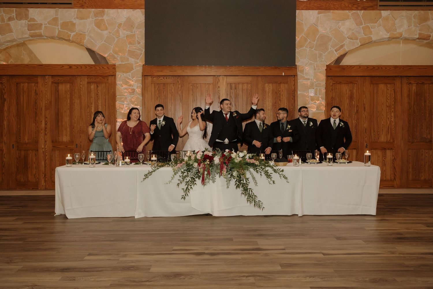 Wedding party raises glasses in a toast at the head table in a rustic stone reception venue.