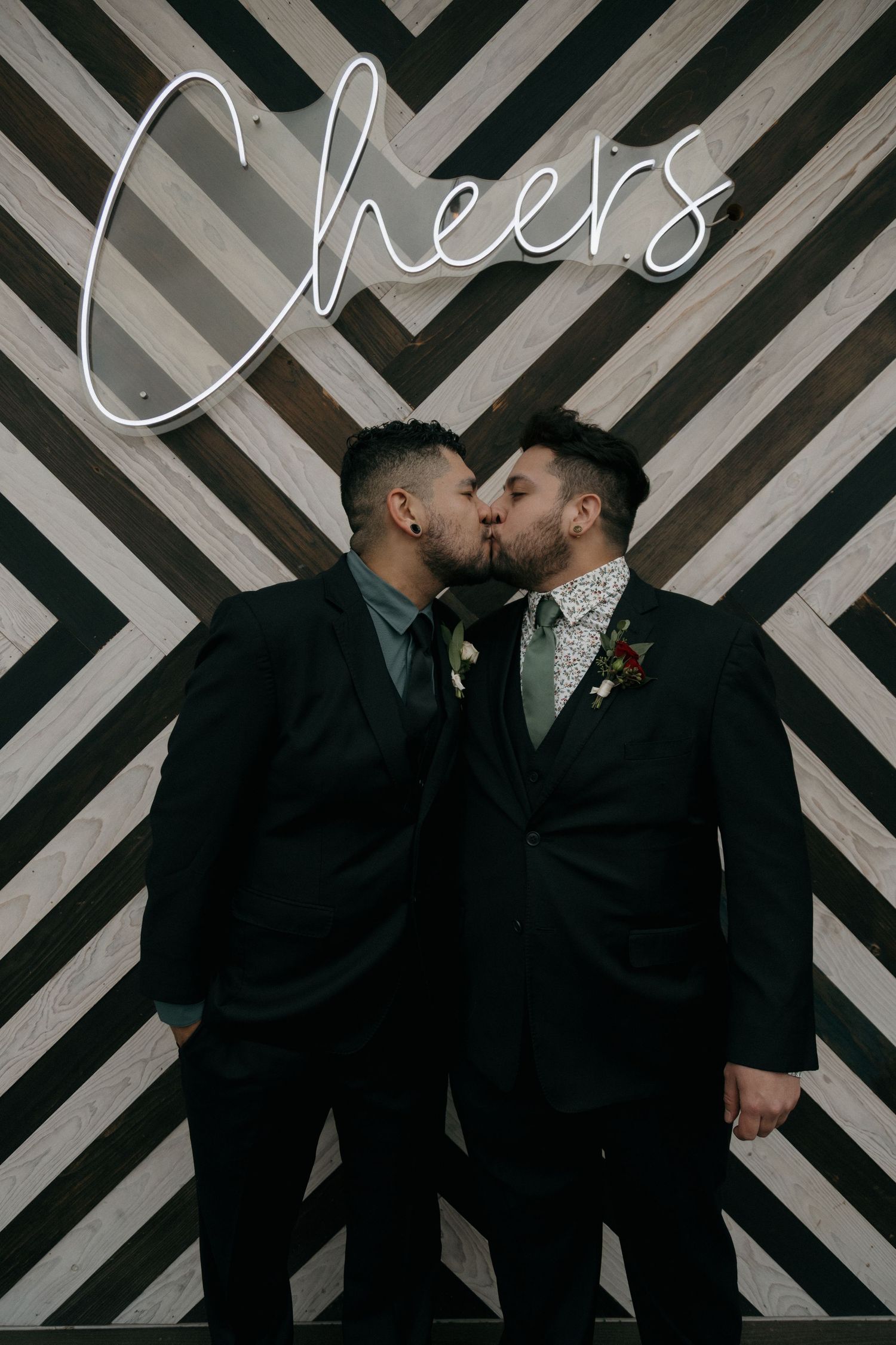 Two grooms share a kiss in front of a black and white striped backdrop with neon Cheers sign.