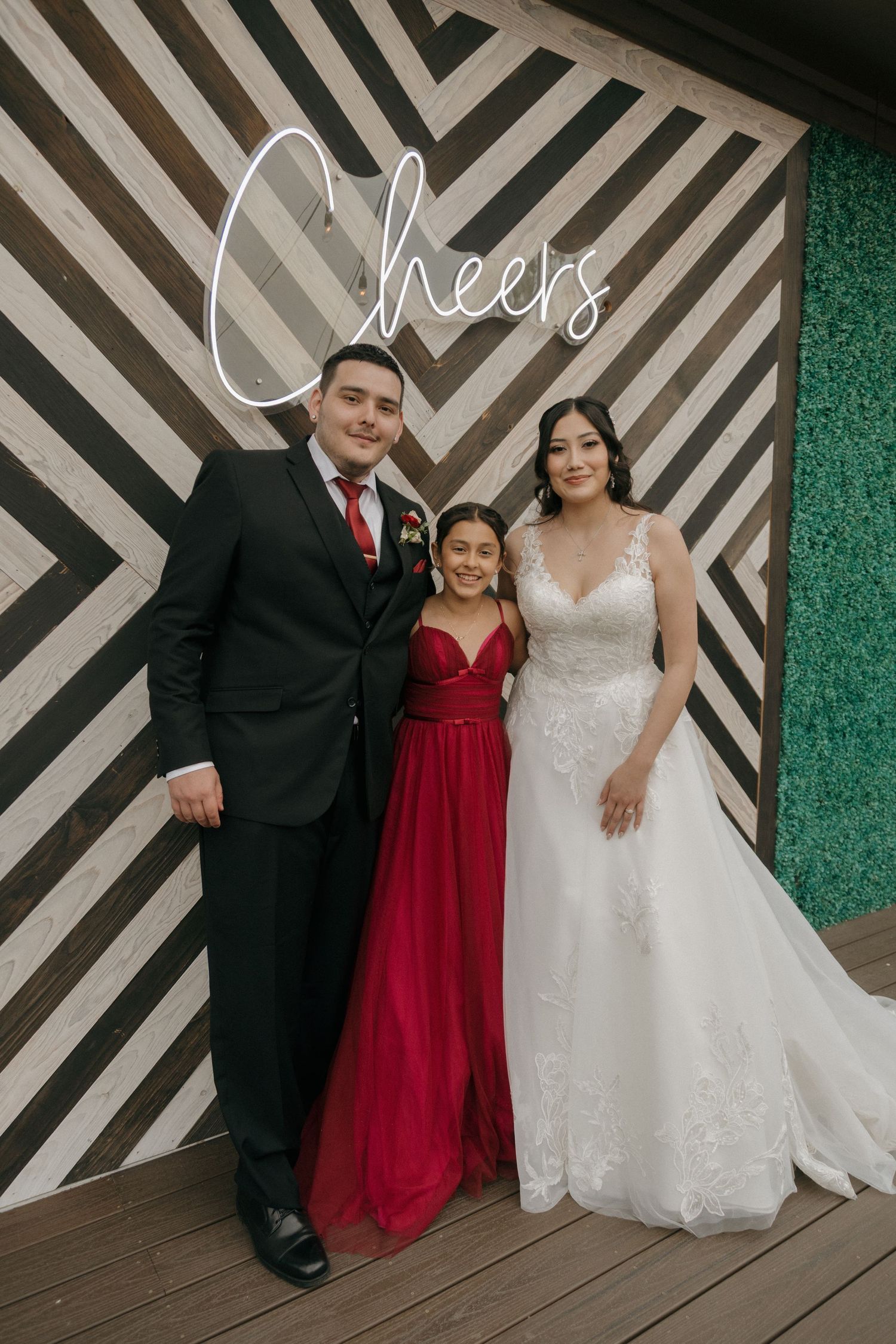 Bride and groom pose with young girl in red dress in front of a geometric chevron backdrop with Cheers neon sign.