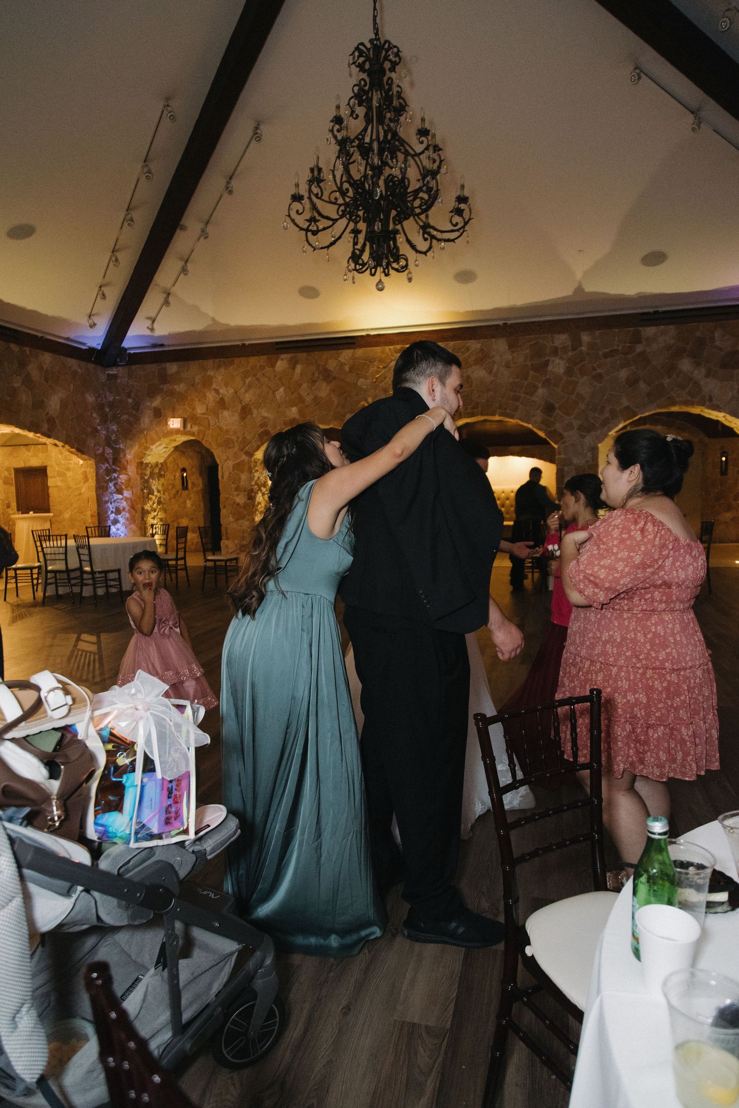 Wedding guests dancing and socializing inside an elegant rustic stone reception hall with chandeliers.