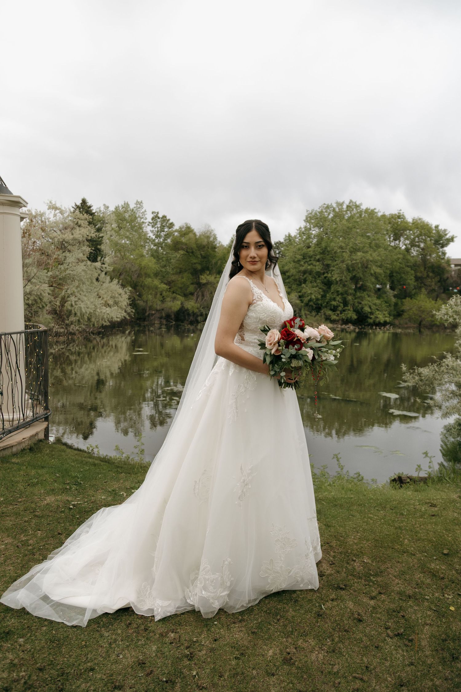 Elegant bride in lace wedding gown holding bouquet of red and pink roses beside a serene pond.
