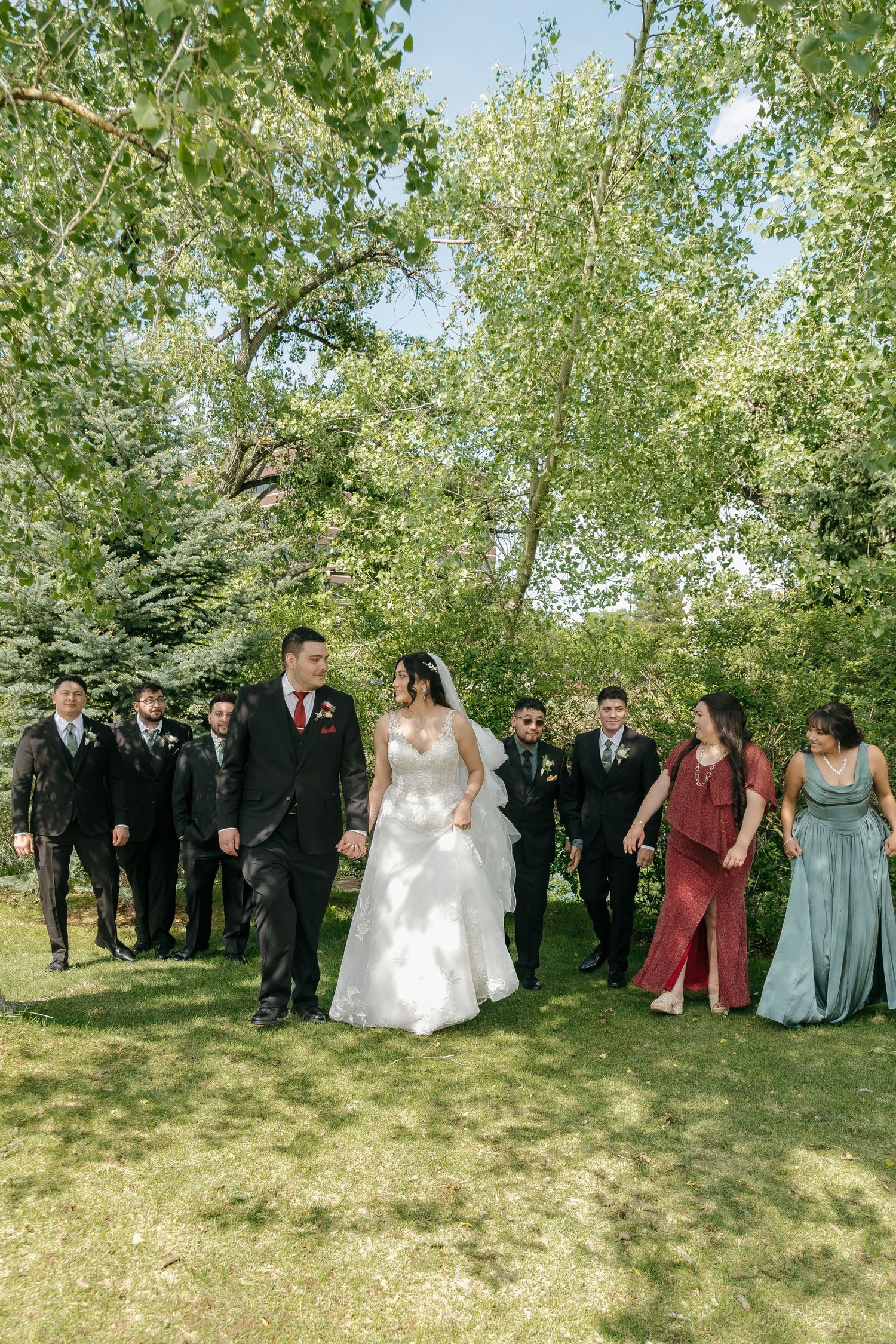Bride and groom walk with wedding party through a sunny green park on wedding day.