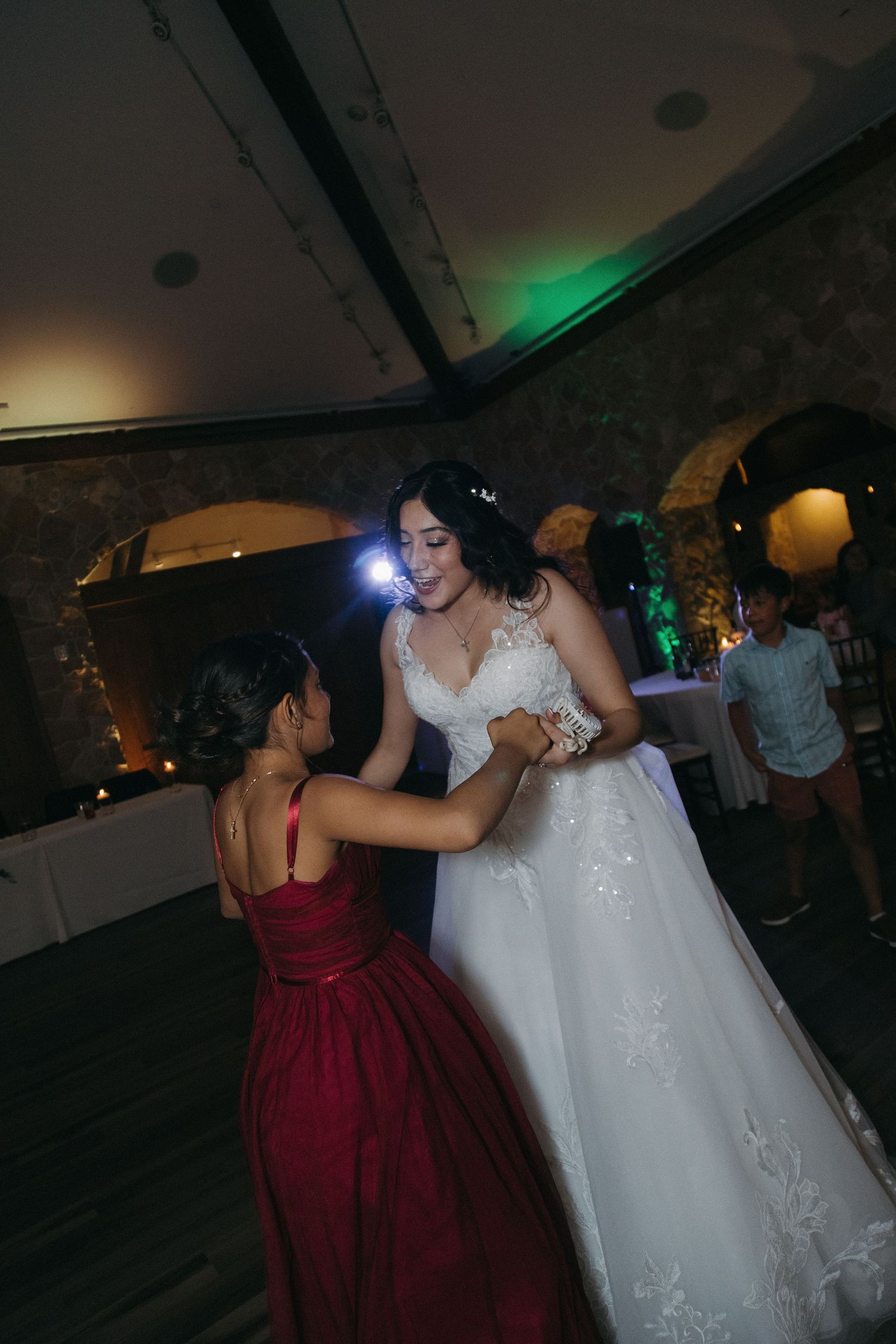 Bride in white lace gown dances with young girl in red dress at wedding reception.