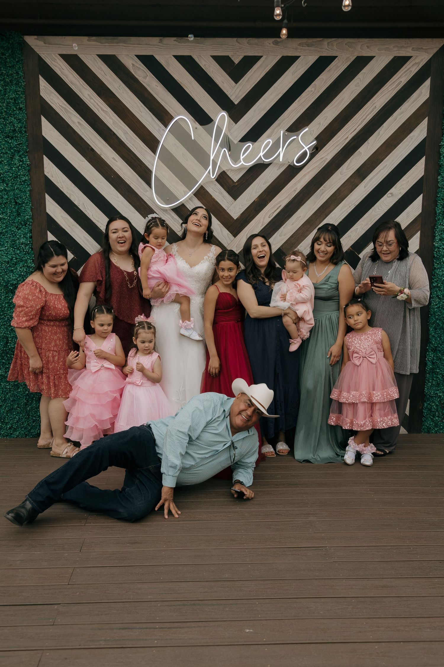 Bride surrounded by female guests and children in front of a Cheers wedding backdrop.