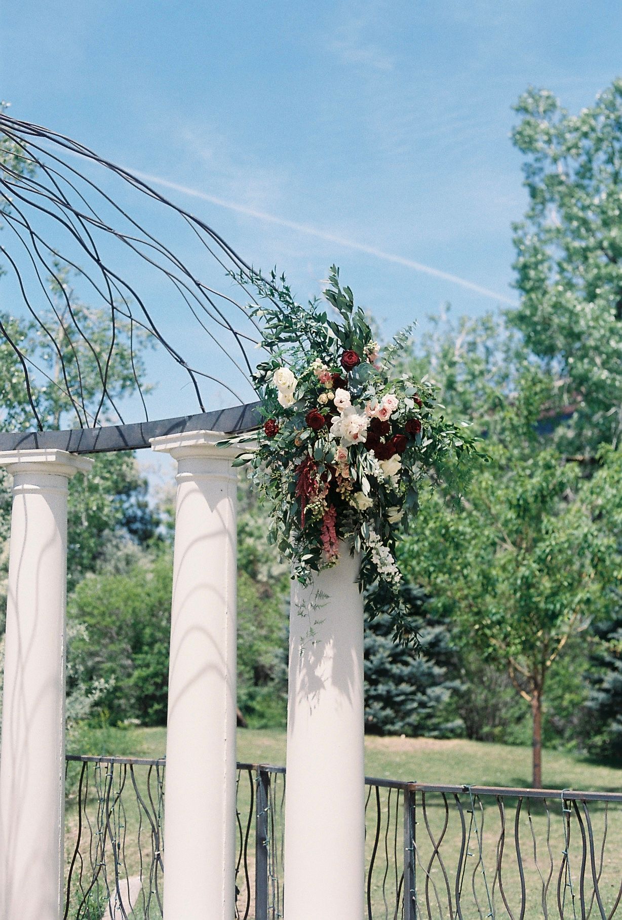 White columns adorned with lush greenery and blush floral arrangement under a decorative iron arch at an outdoor wedding venue.