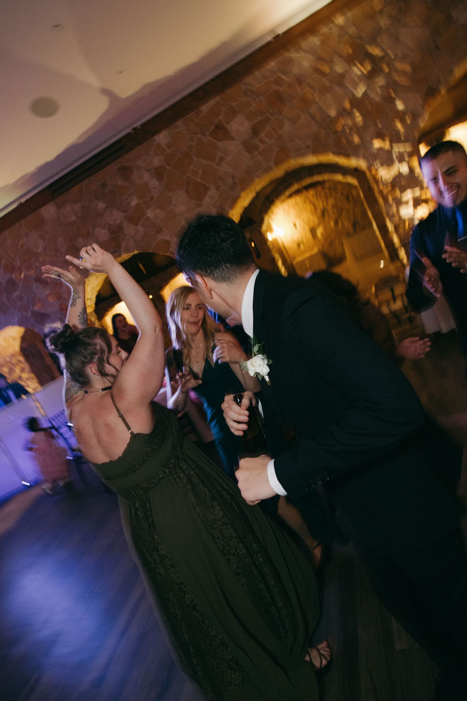 Groom in black suit dances energetically with bridesmaid in olive dress at lively wedding reception.