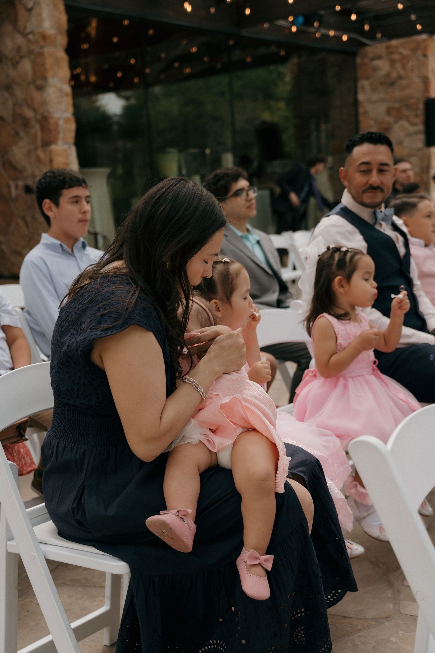 Woman in navy dress holds a baby in pink at an outdoor wedding ceremony with guests seated nearby.