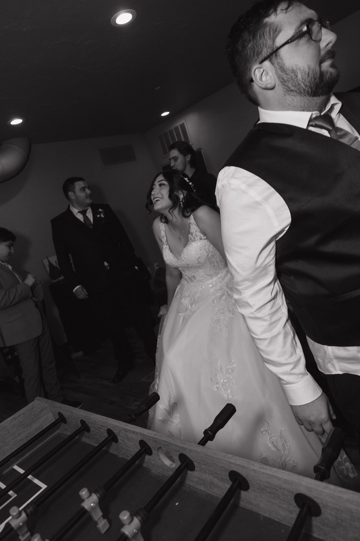 Bride in lace gown playing foosball with guests at wedding reception.