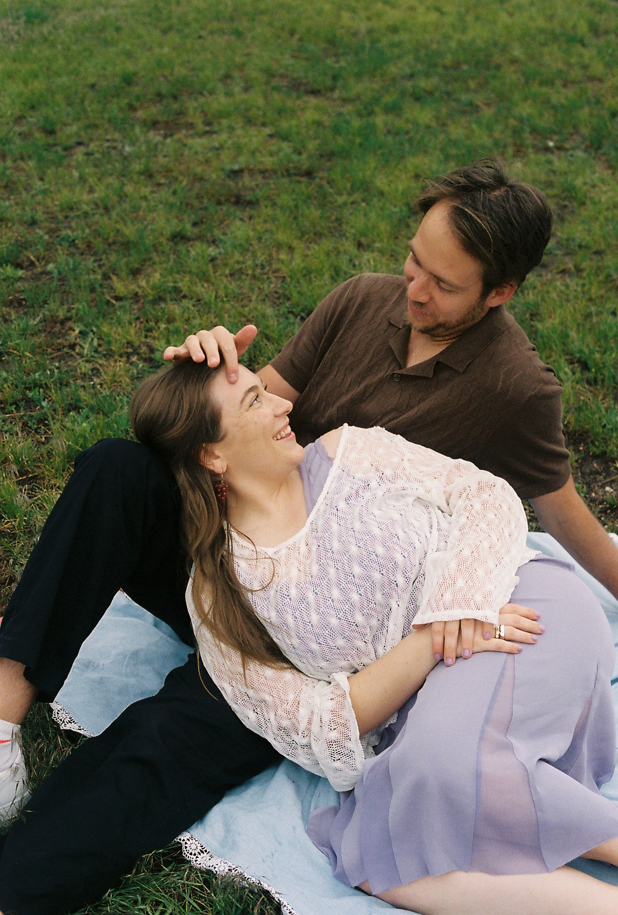 Couple laughing and relaxing together on a blanket in a lush green park during a picnic.
