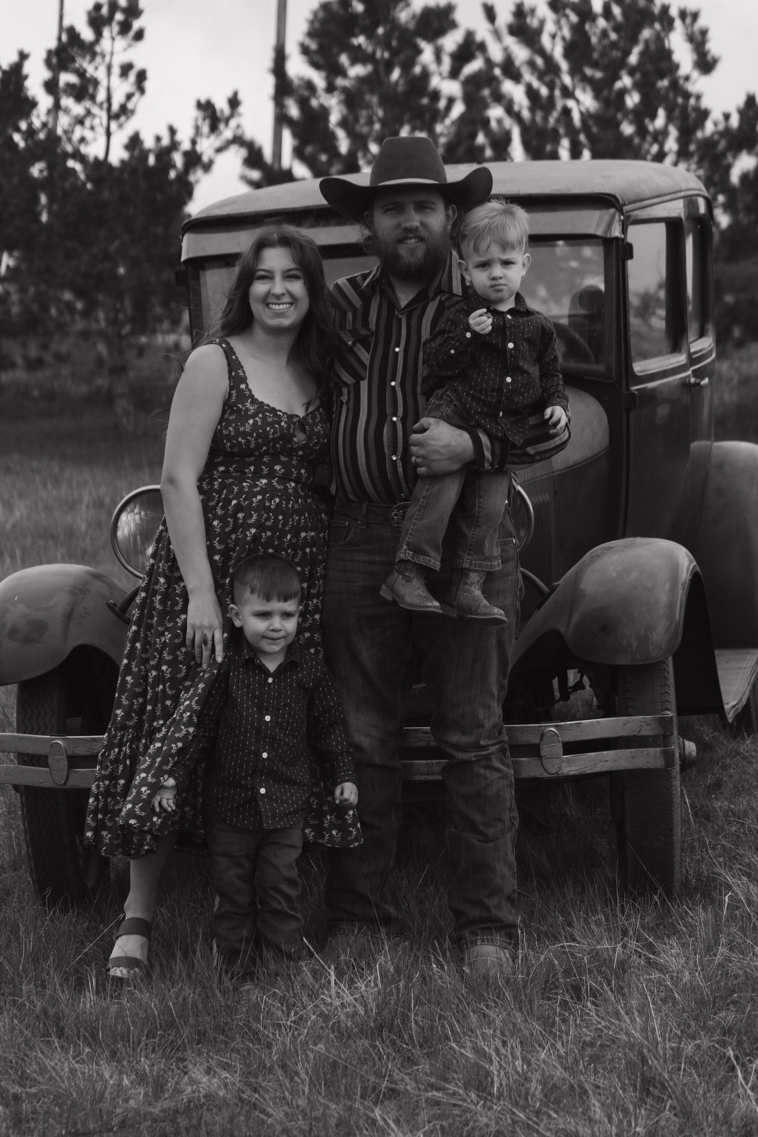 Black and white family portrait with rustic vintage car backdrop.