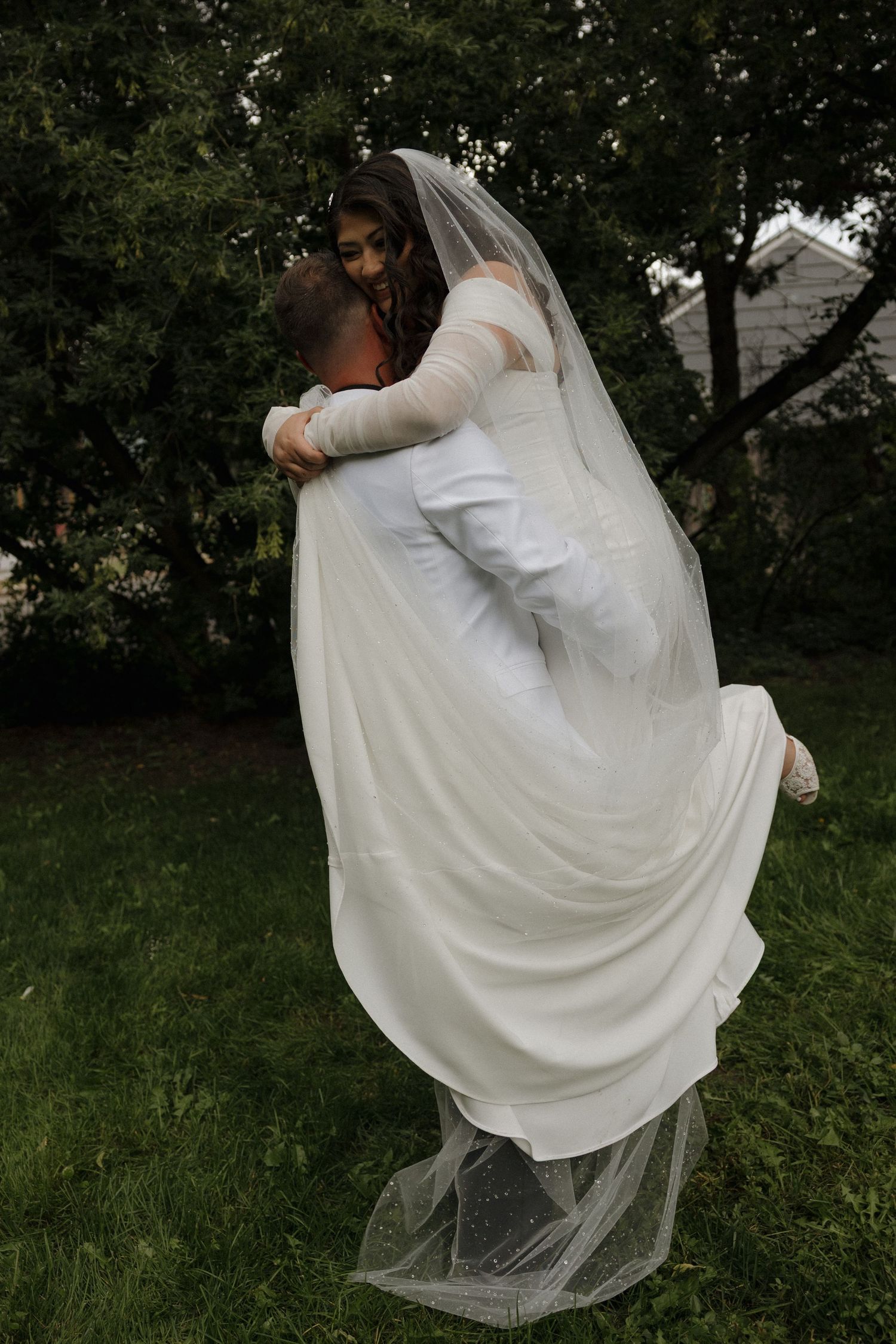 Groom lifting bride in white wedding dress and veil outdoors on green grass with trees in background.