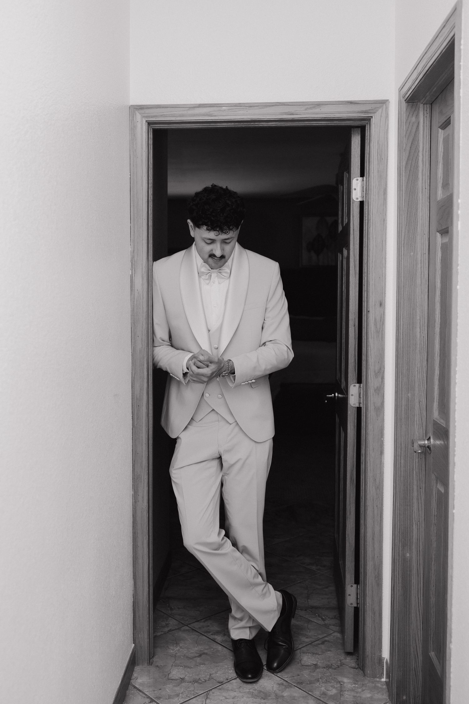 Groom in light suit and bow tie adjusting cufflinks in doorway, black and white wedding portrait.