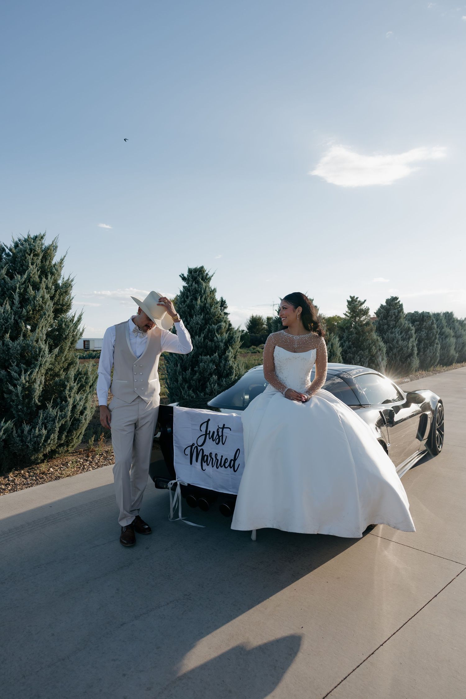 Bride and groom pose beside a sports car with a Just Married sign on their wedding day outdoors.