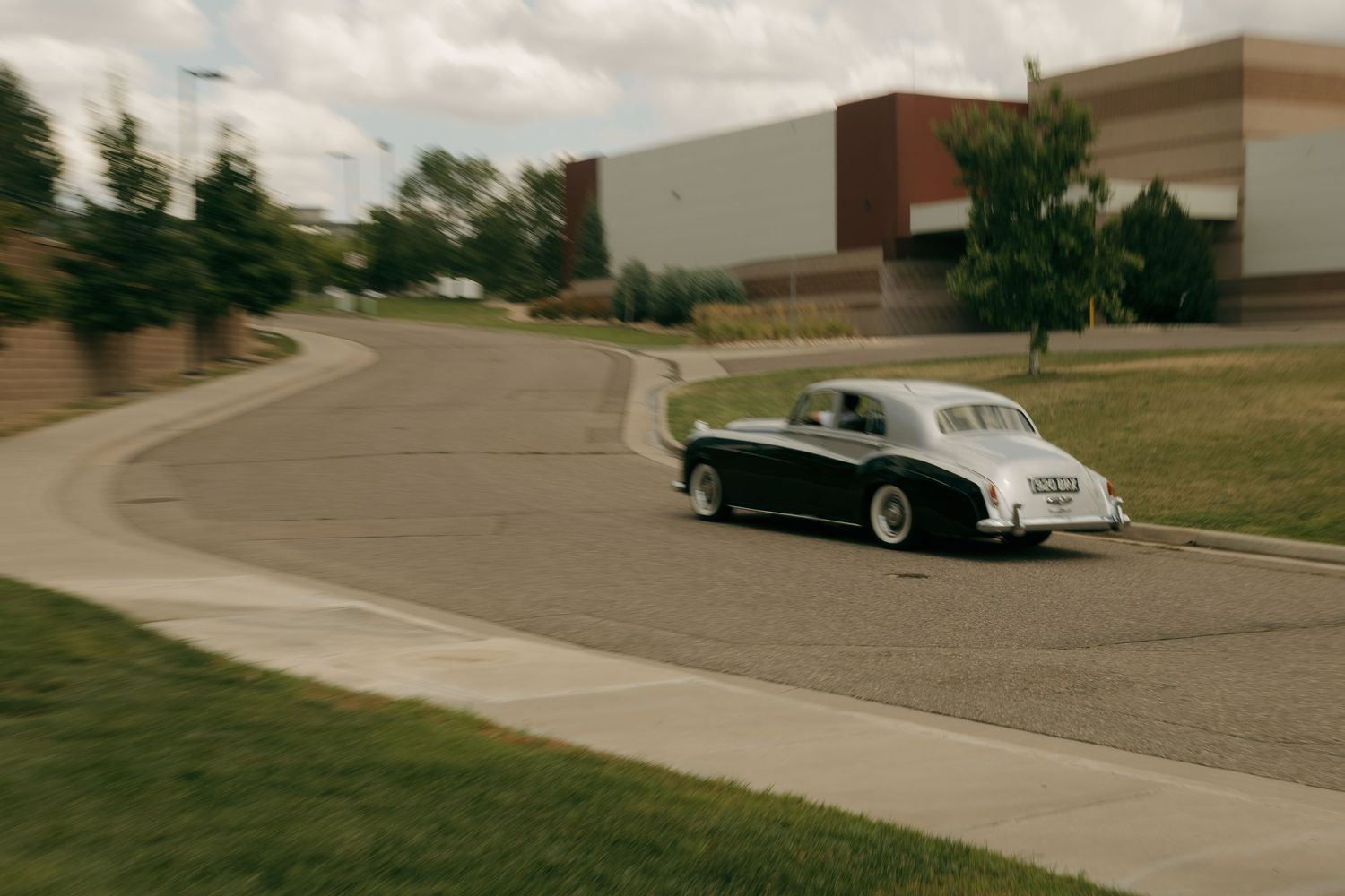 A classic vintage black and white car drives along a curved road near a modern brick building.