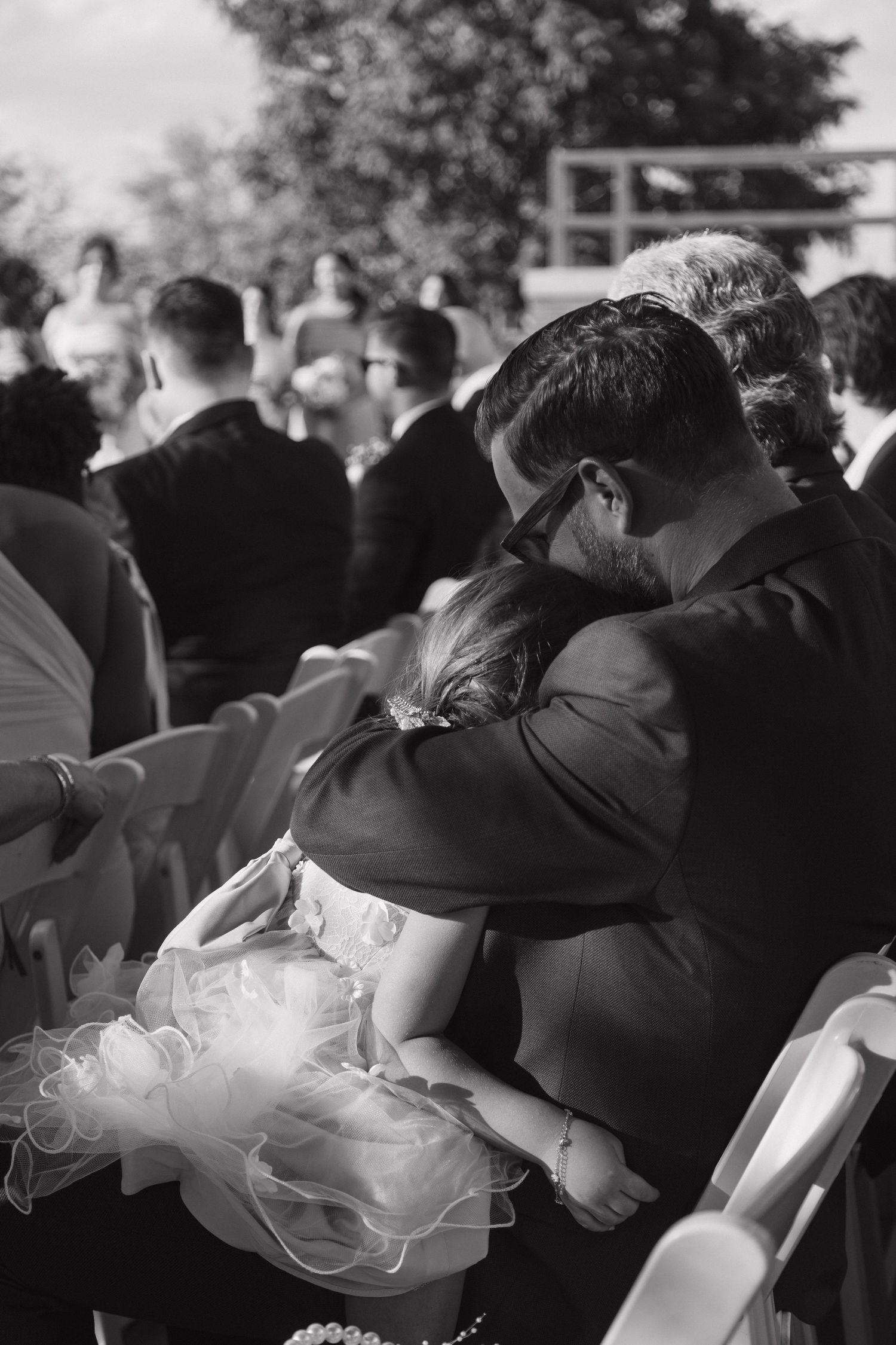Man in suit embraces young girl in white dress at outdoor wedding ceremony, black and white photo.