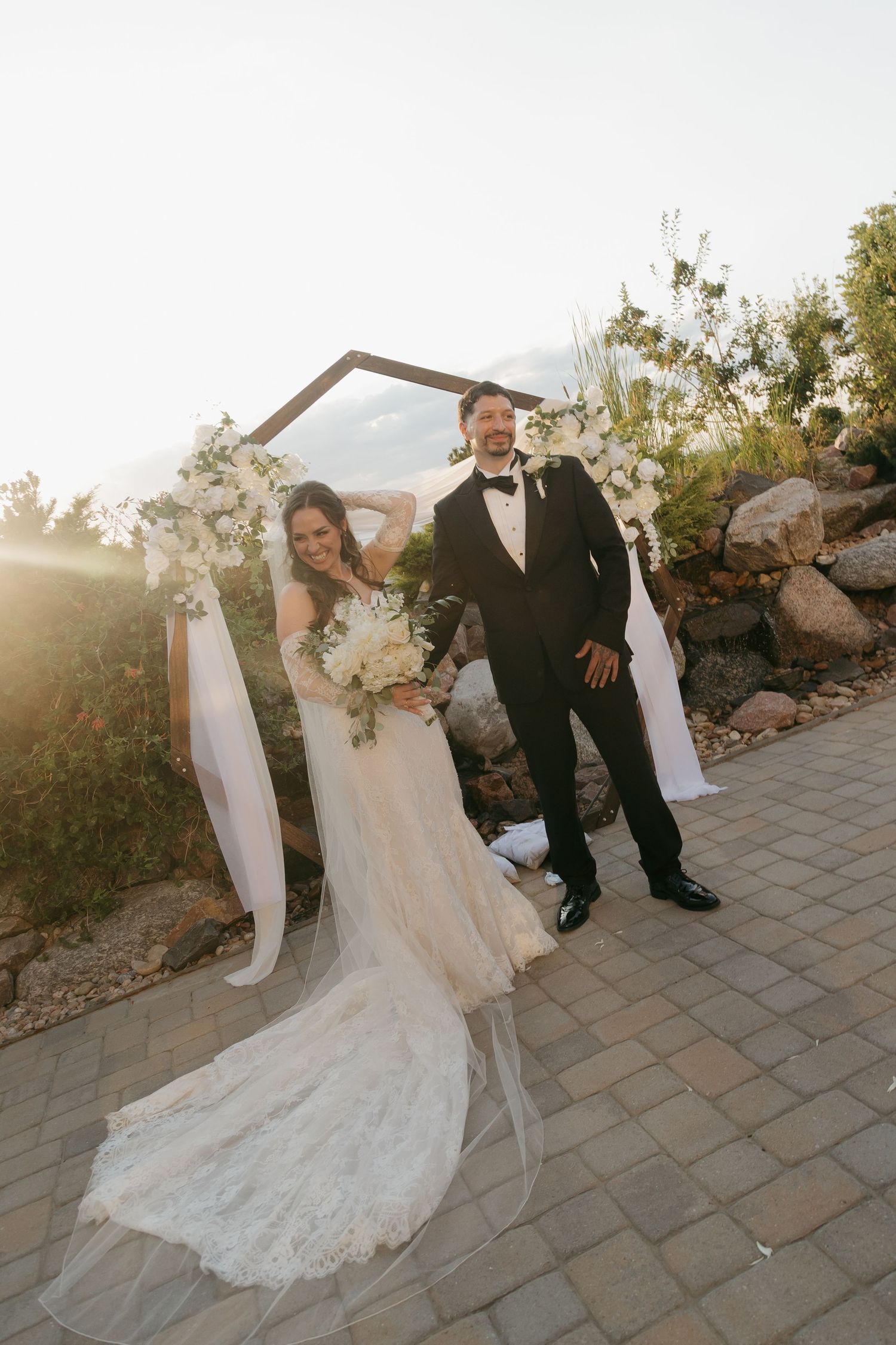 Bride and groom posing under floral arch at golden hour outdoor wedding ceremony.
