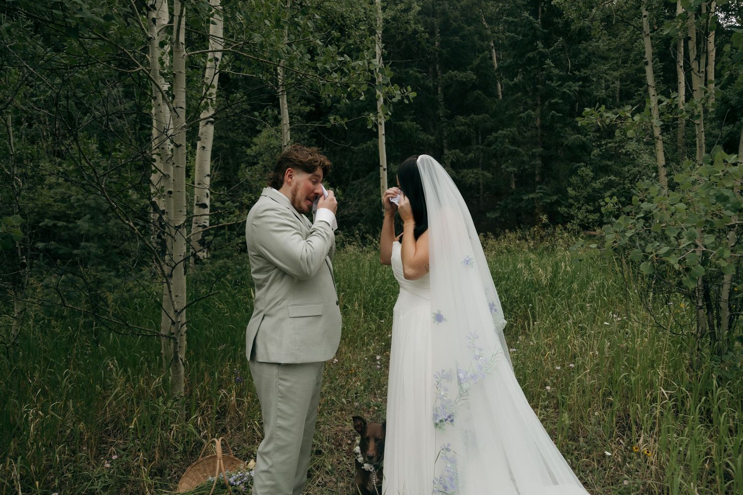 Emotional groom wiping tears during first look with bride in white gown and veil in lush forest wedding setting.