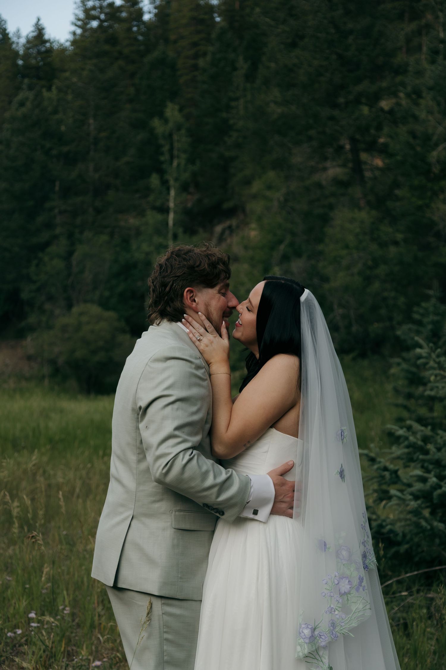 Bride and groom share a romantic kiss surrounded by tall evergreen trees in a forest meadow.