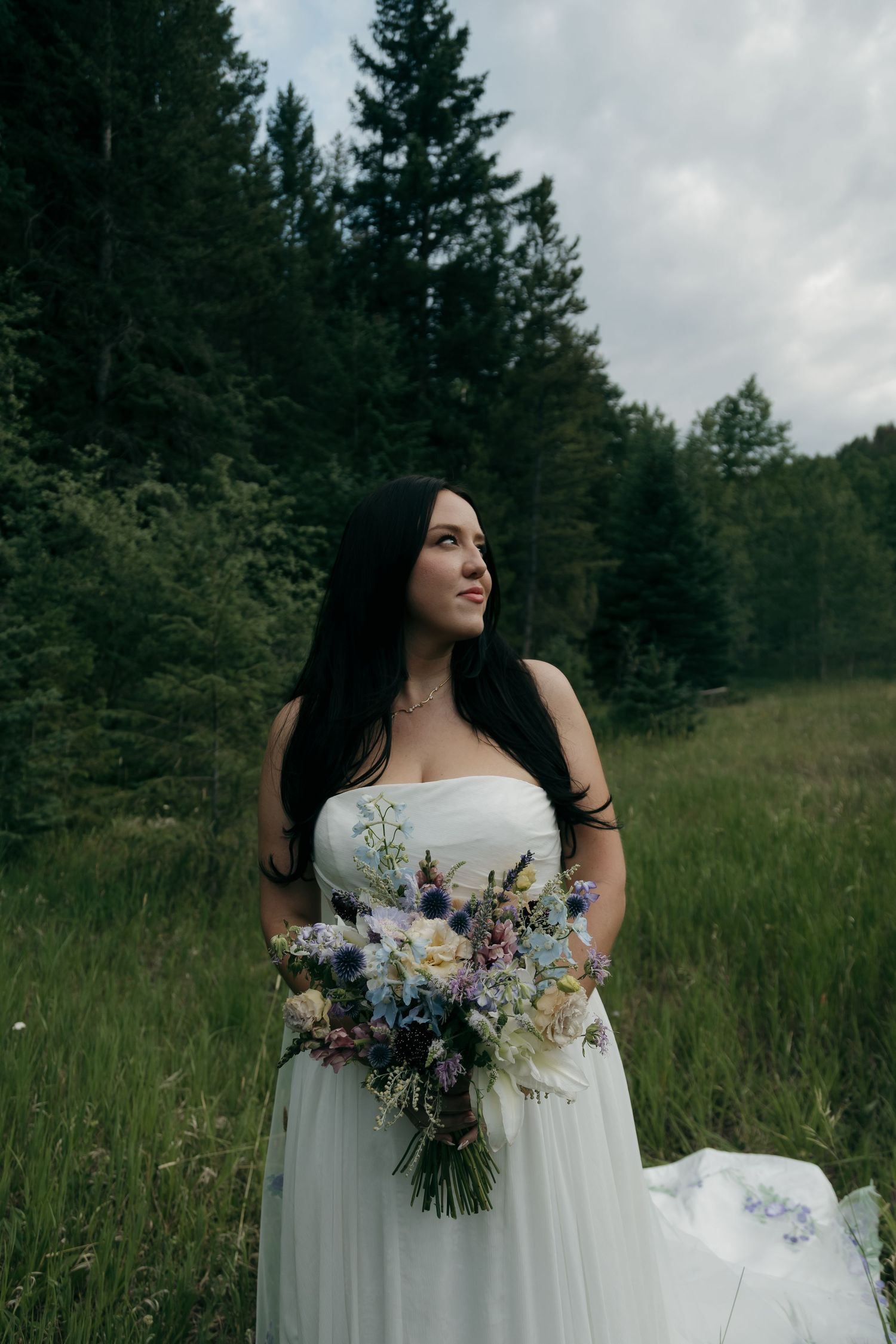 Dark-haired bride in a strapless white gown holds a wildflower bouquet in a forest meadow setting.