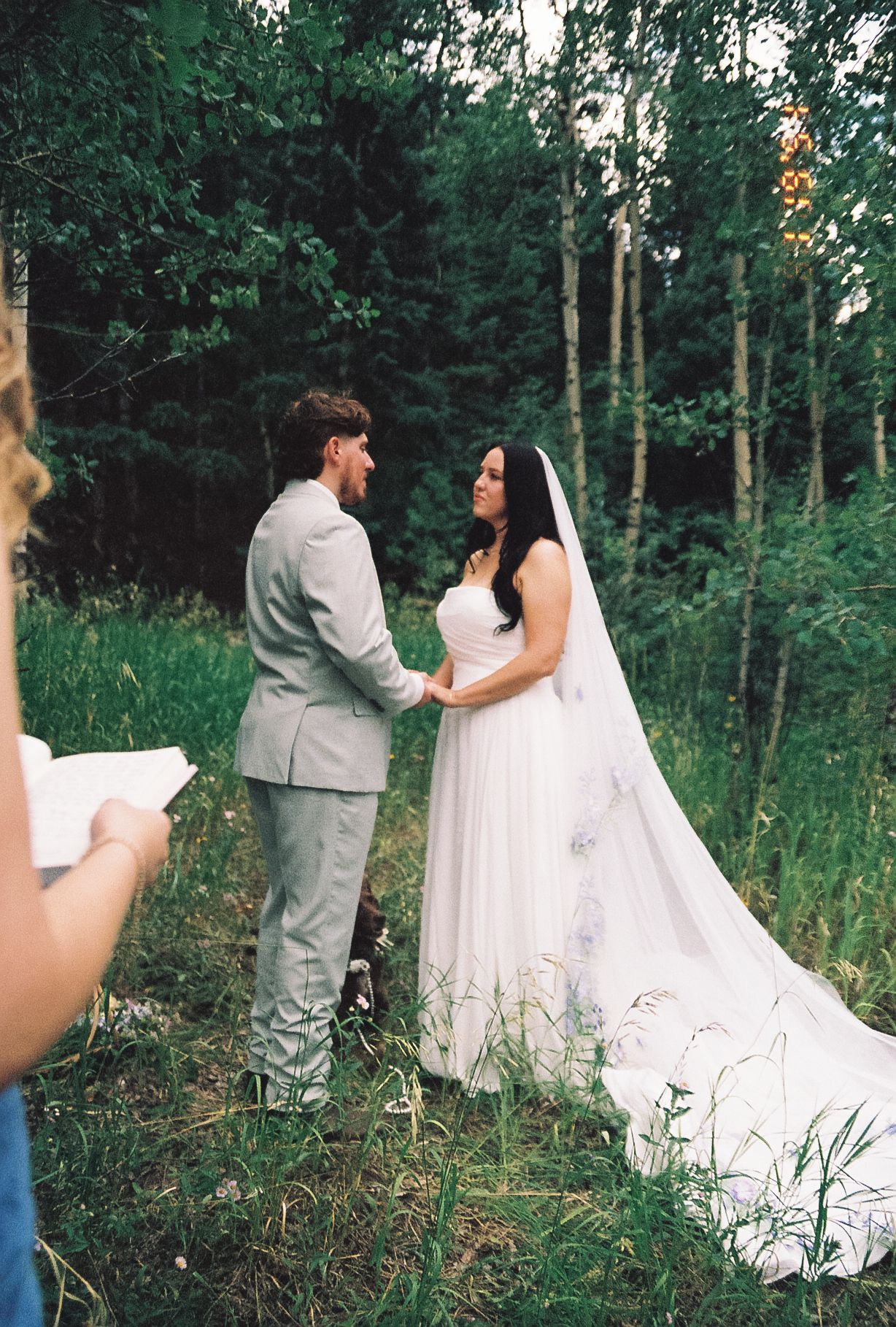 Bride in white gown with veil and groom in gray suit exchanging vows in a lush forest clearing.