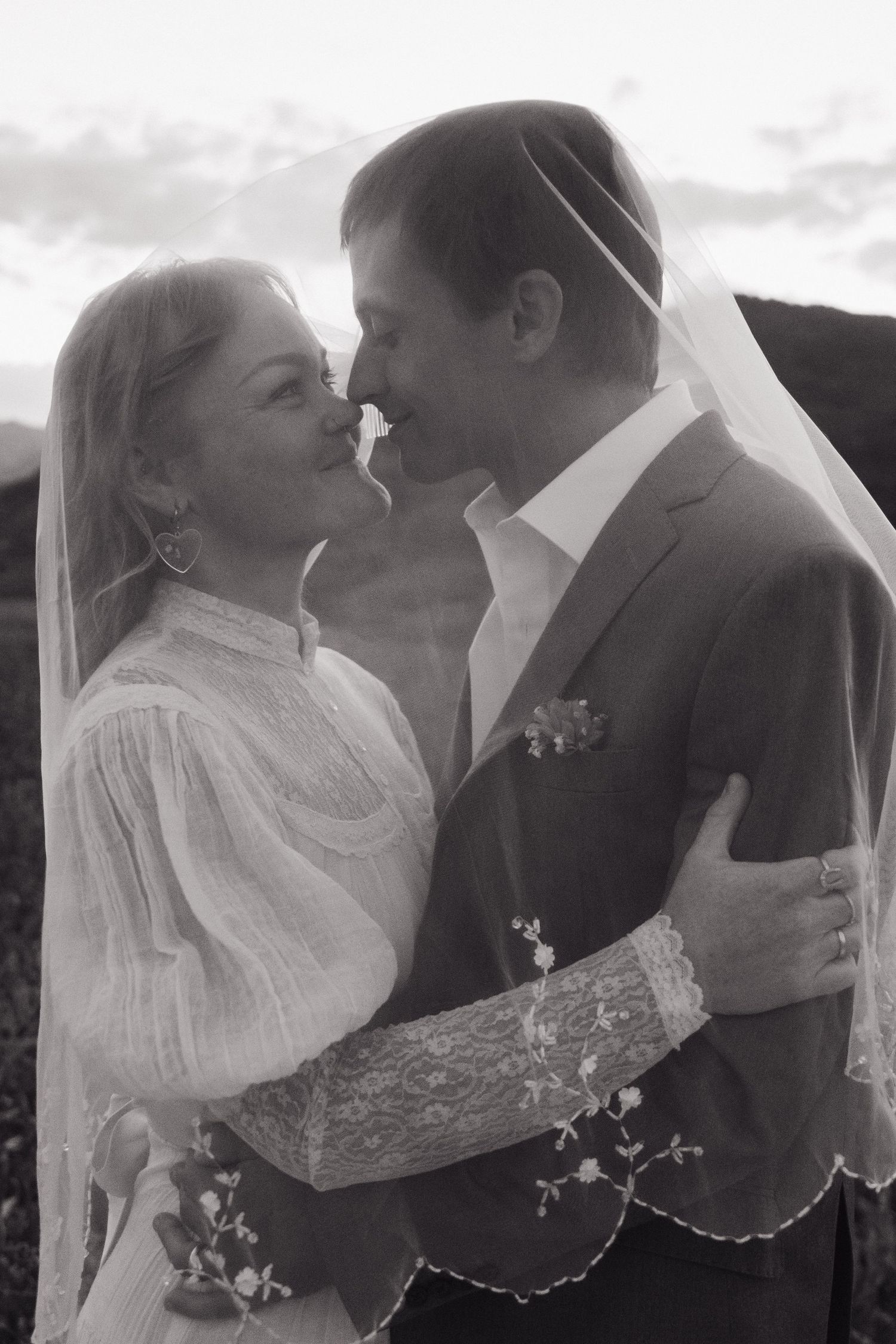 Black and white photo of bride and groom sharing a tender kiss under a lace wedding veil outdoors.