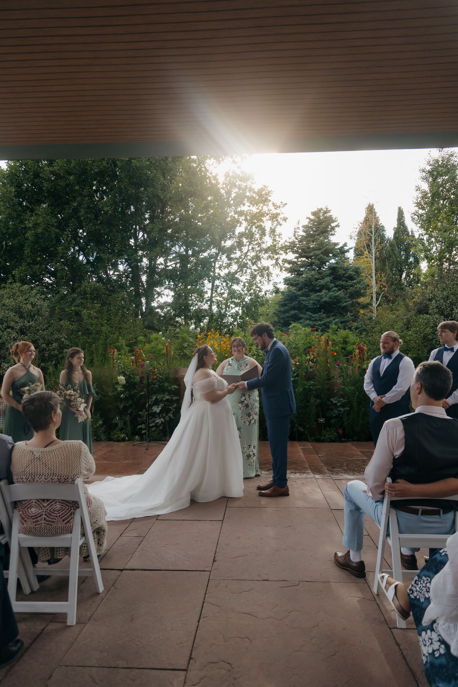 Wedding ceremony on sunlit outdoor patio with bride in white gown and groom in navy suit.