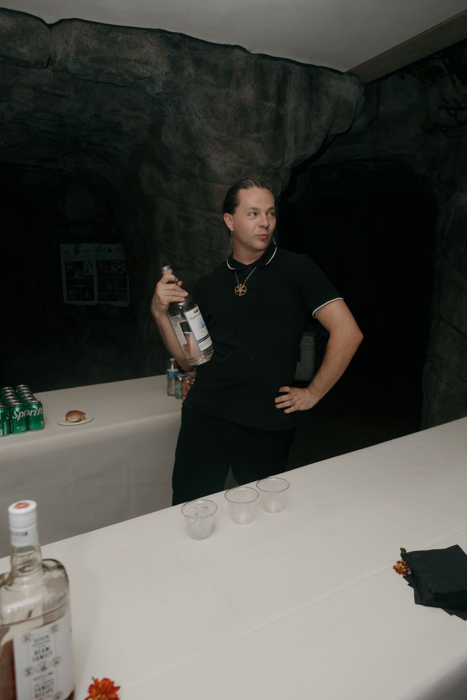 Bartender in black polo holds liquor bottle at cave-themed wedding bar setup.