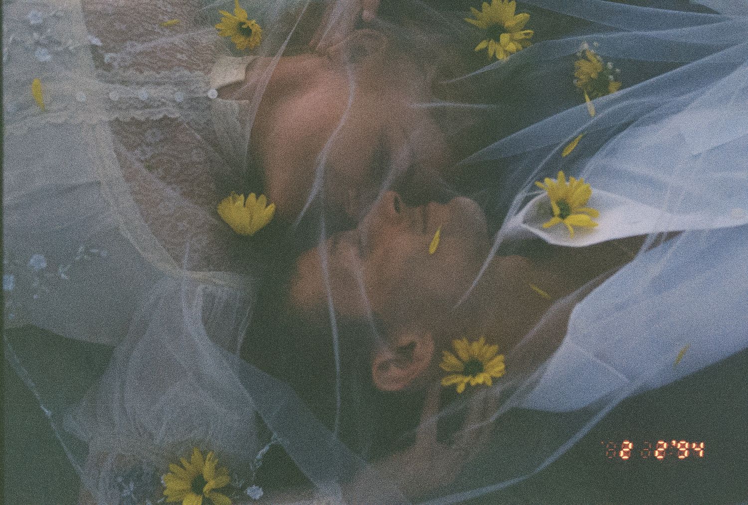 Romantic couple kissing under a sheer bridal veil adorned with yellow daisies in a dreamy film photo.