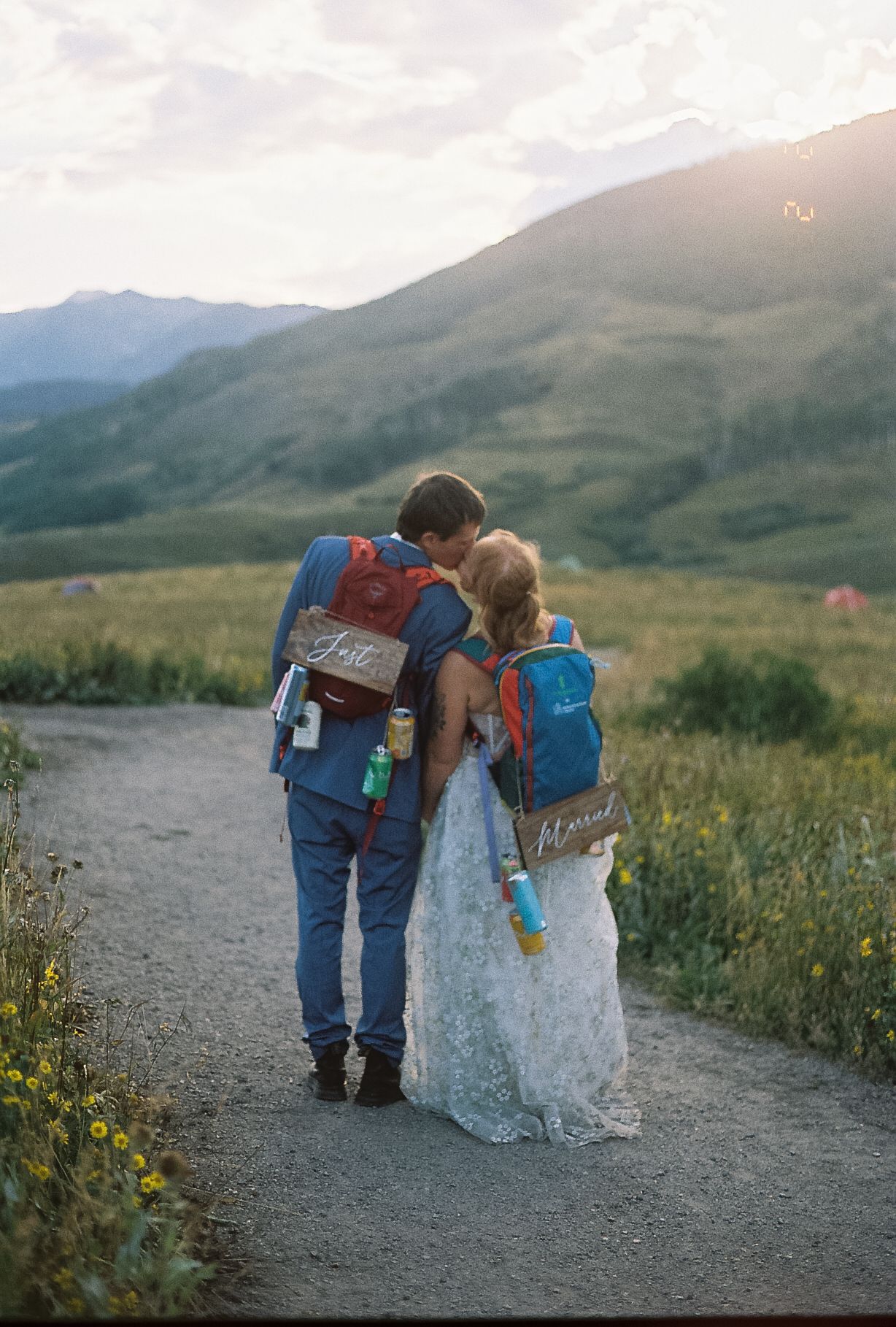 Newlyweds walking away with backpacks labeled Just Married on a mountain path at sunset.