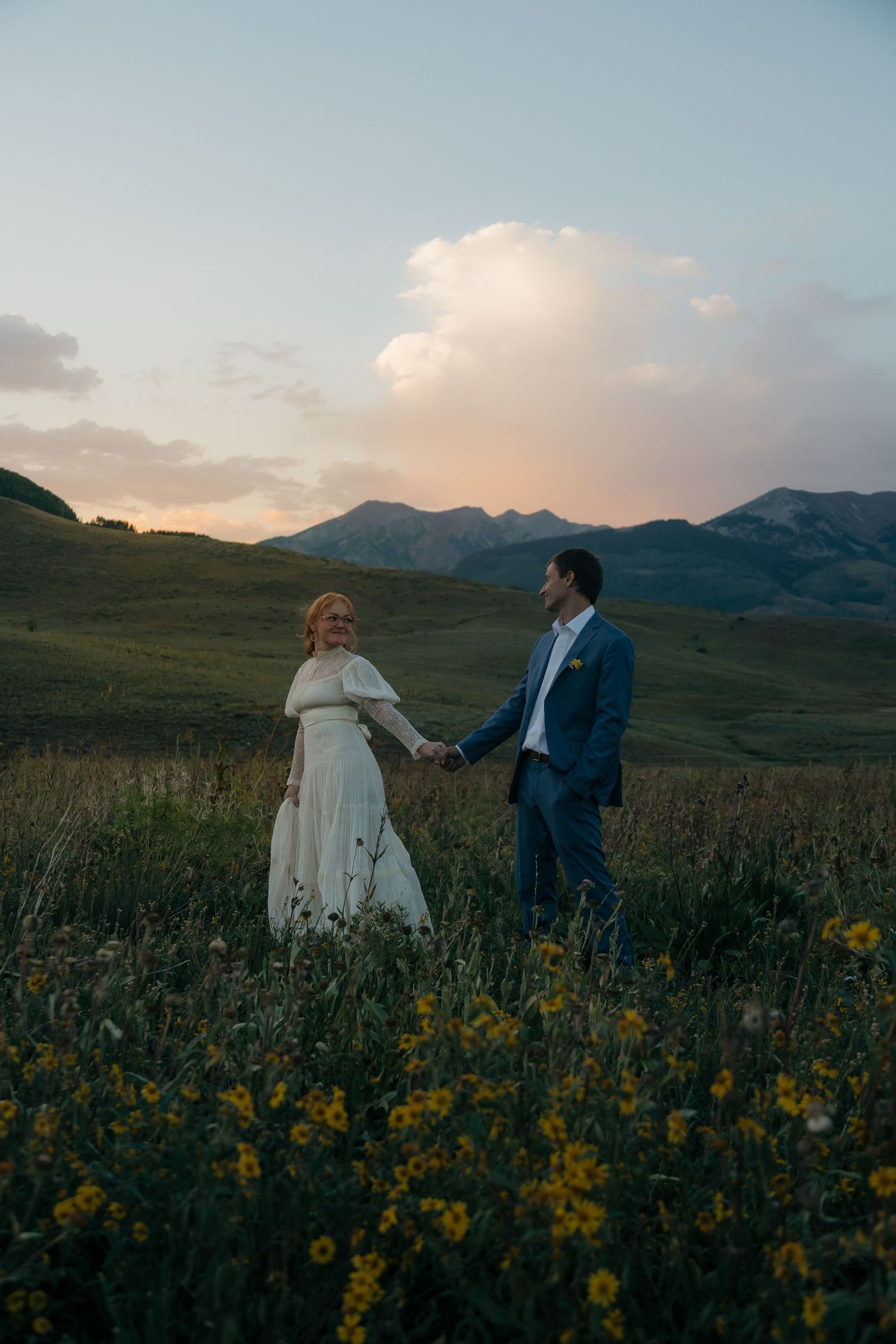 Newlywed couple holds hands walking through yellow wildflower field with stunning mountain backdrop at sunset.