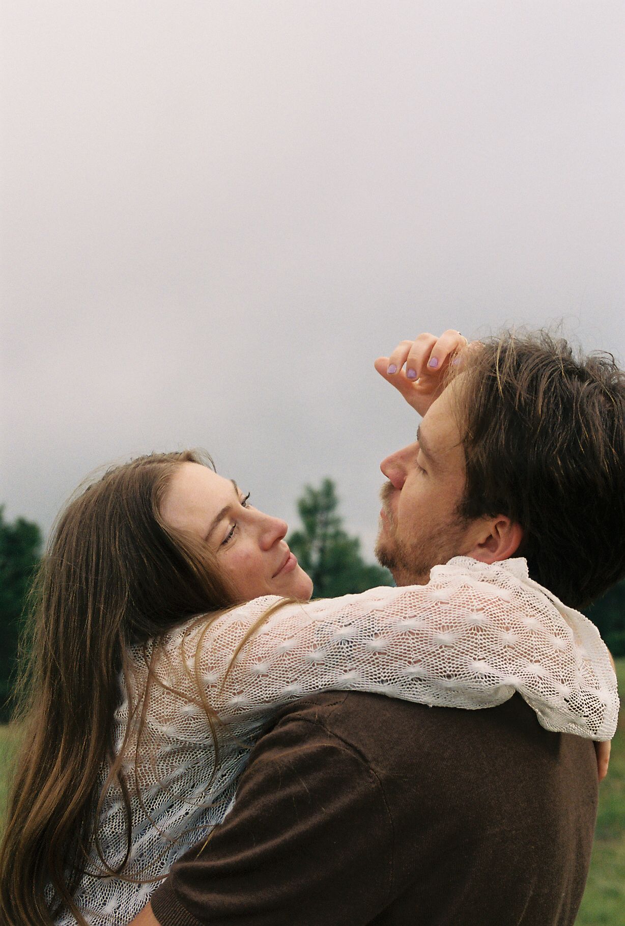 Romantic couple embracing closely outdoors, woman in white lace top resting against man in brown shirt.