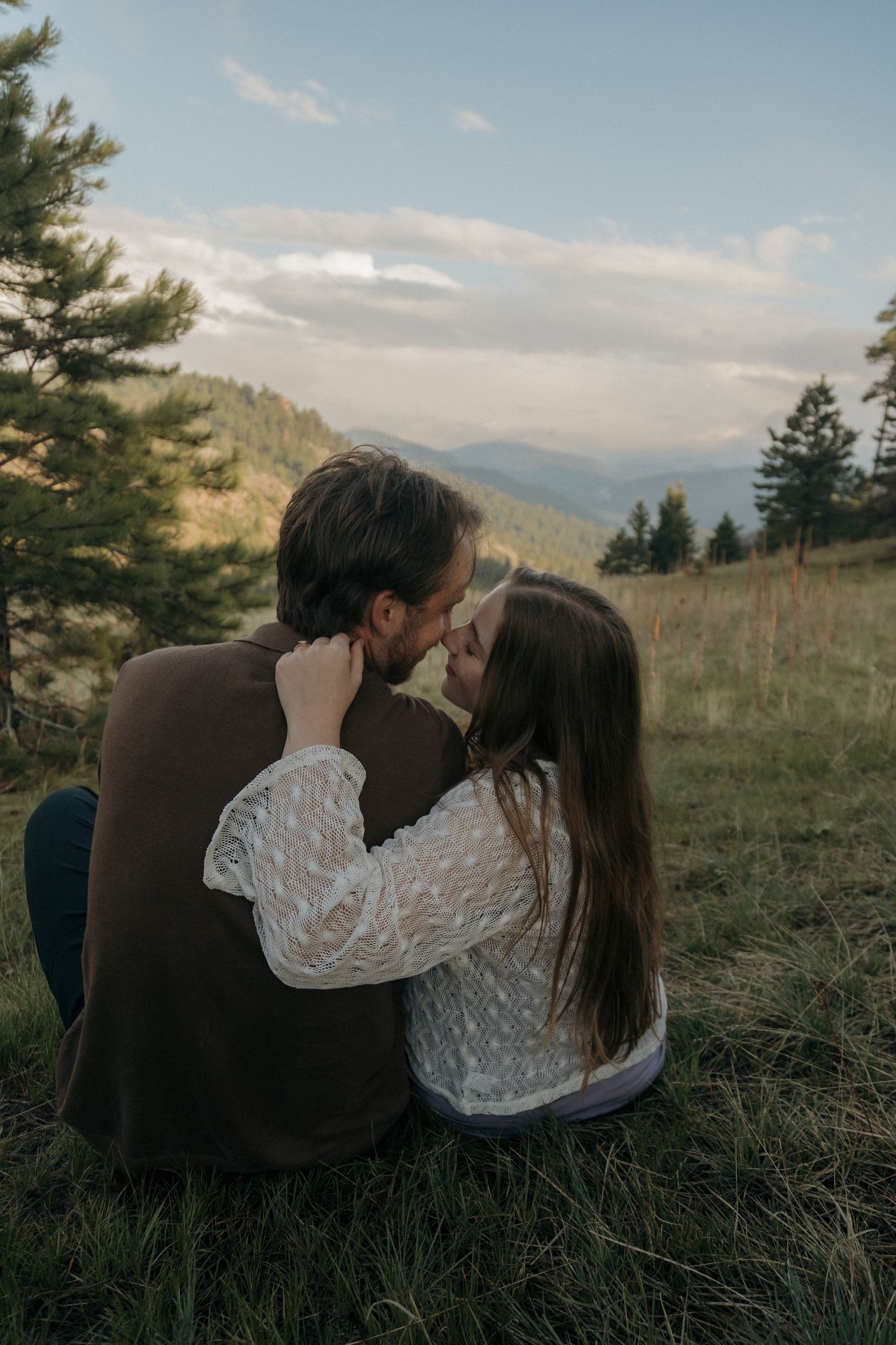 Couple sharing a romantic moment outdoors on a mountain meadow surrounded by pine trees at sunset.