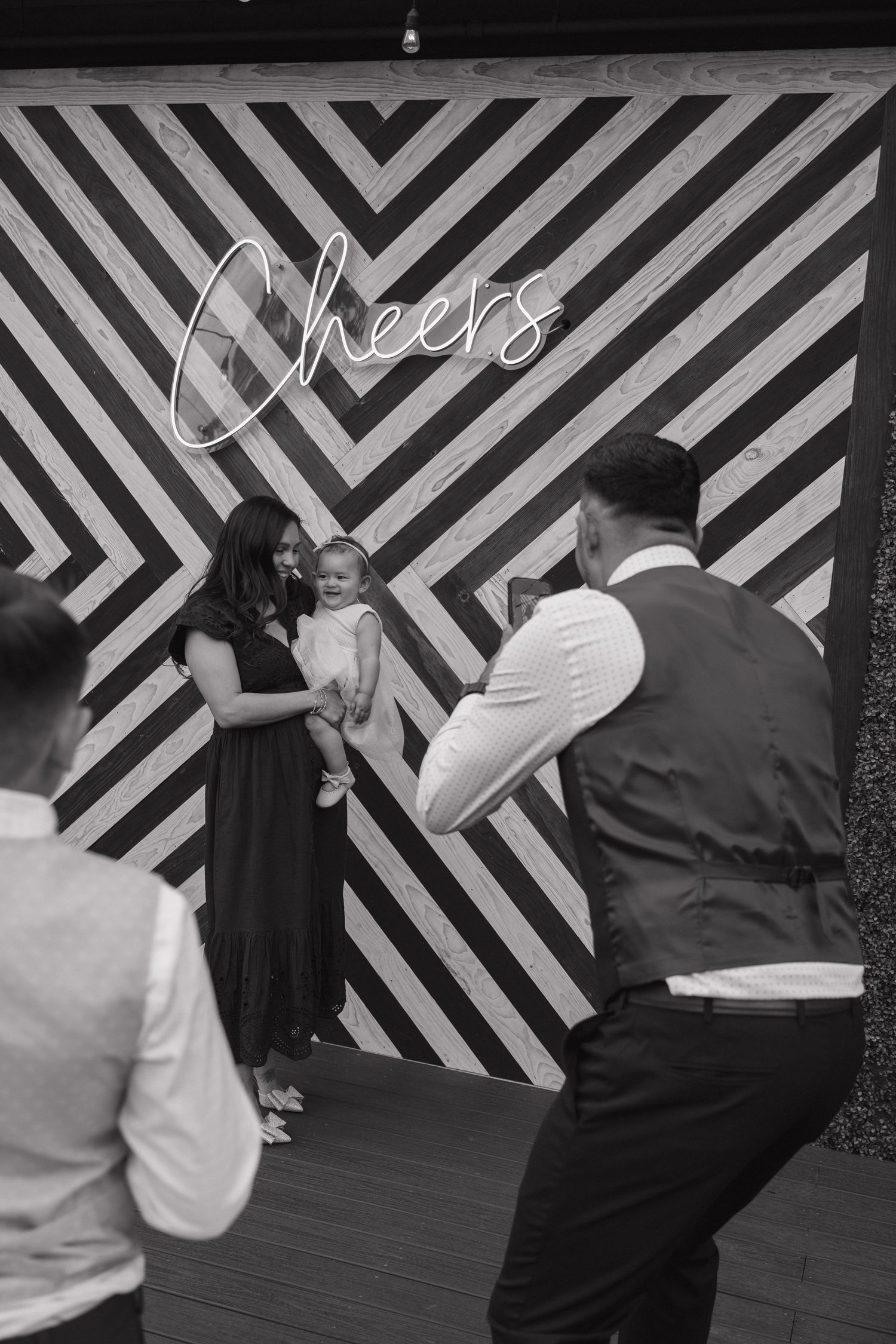 Black and white photo of woman holding baby posing at Cheers wedding backdrop.