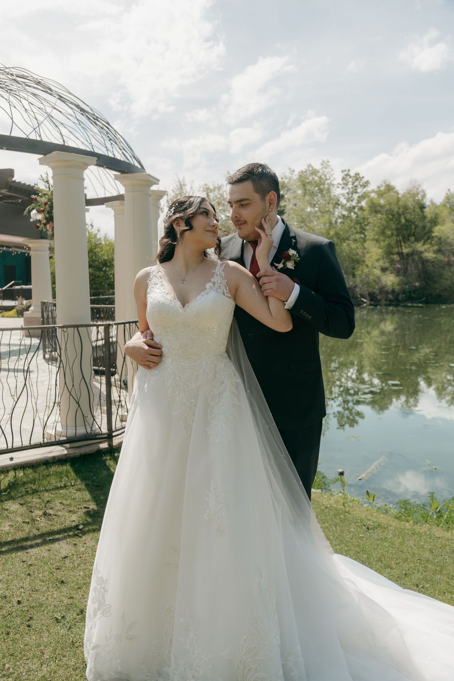 Bride and groom sharing a tender moment near a white gazebo by a lake on their wedding day.