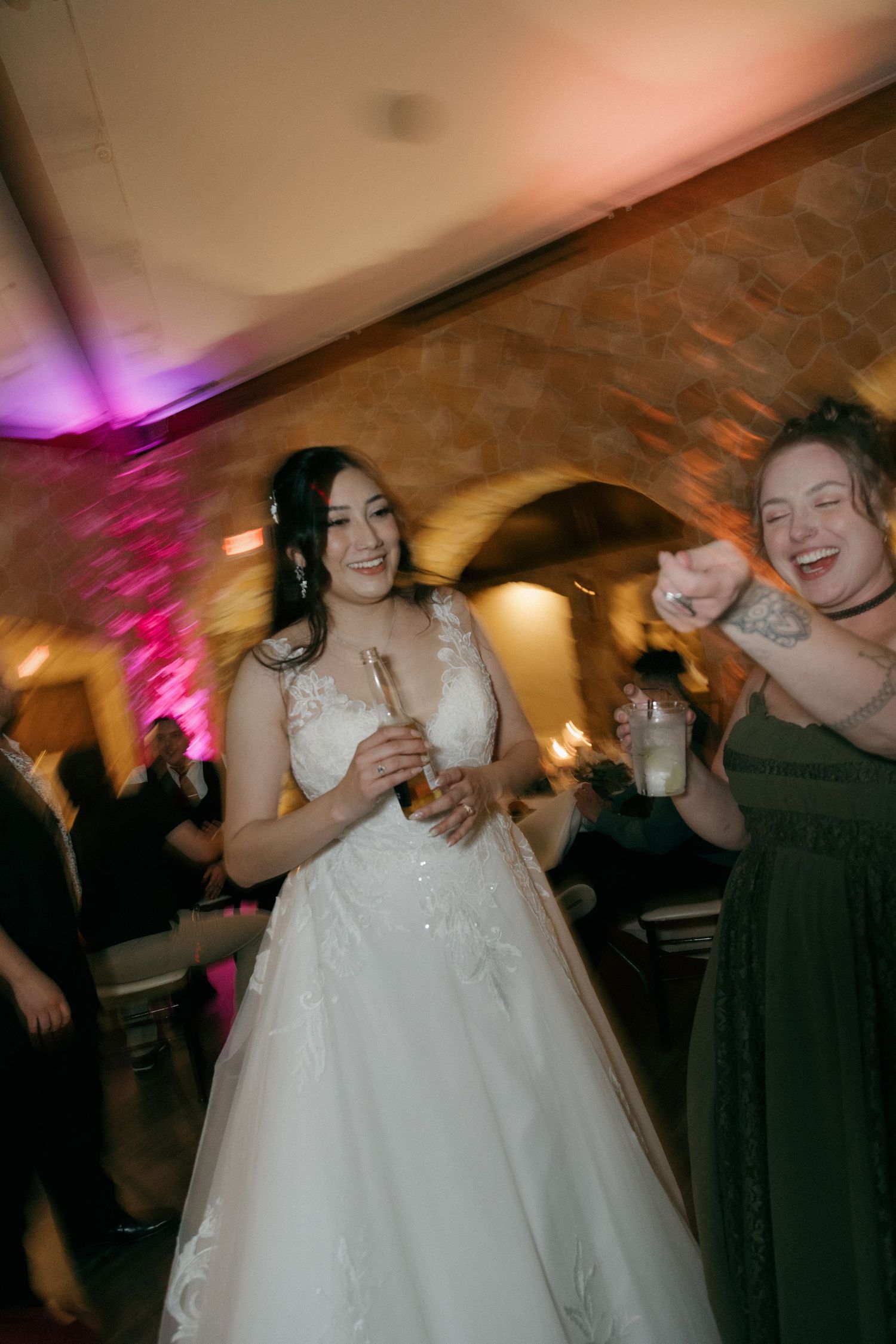 Smiling bride in white lace gown dances joyfully with a friend at a warmly lit wedding reception.