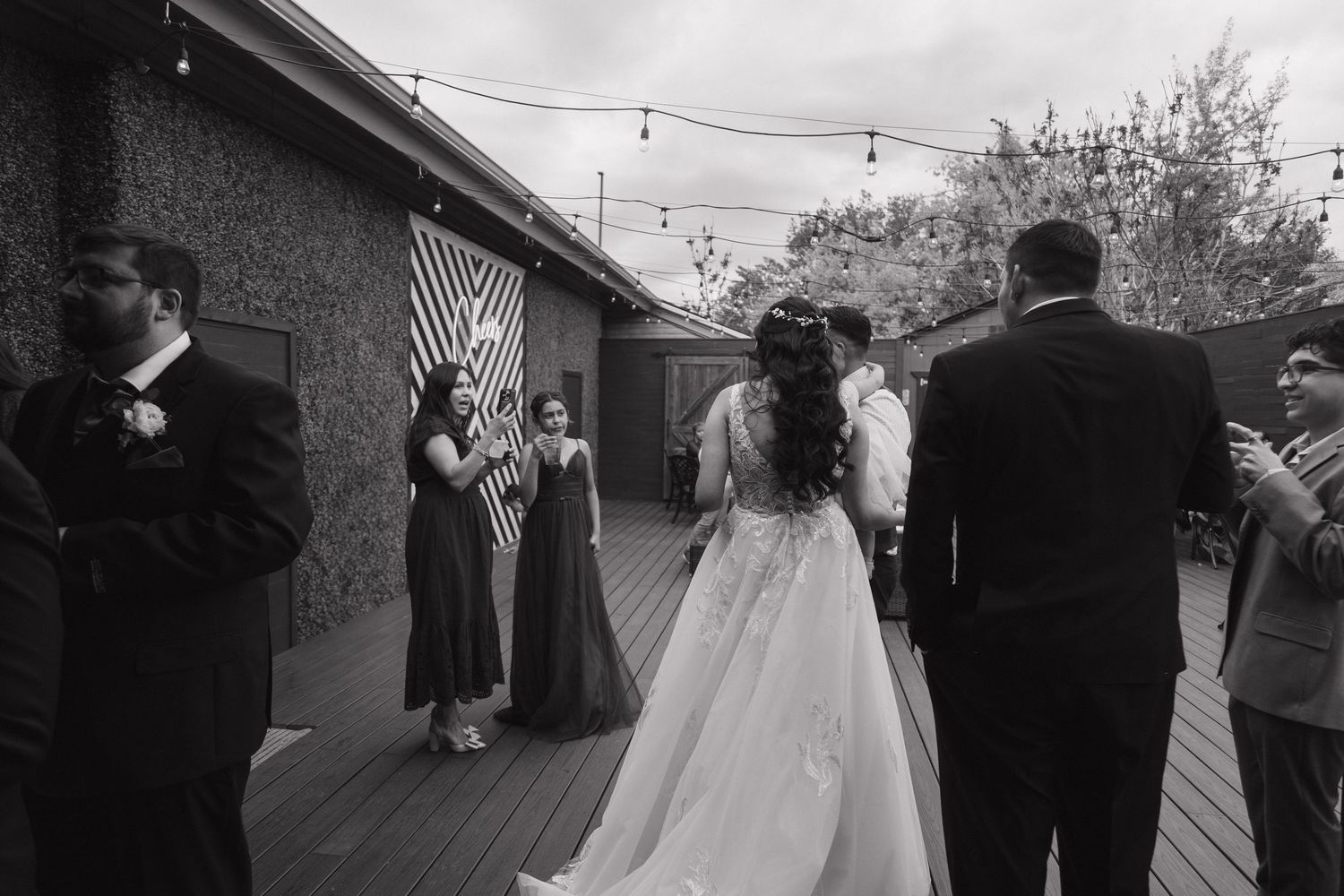 Bride in lace gown walks toward wedding party gathered on outdoor deck with string lights above.