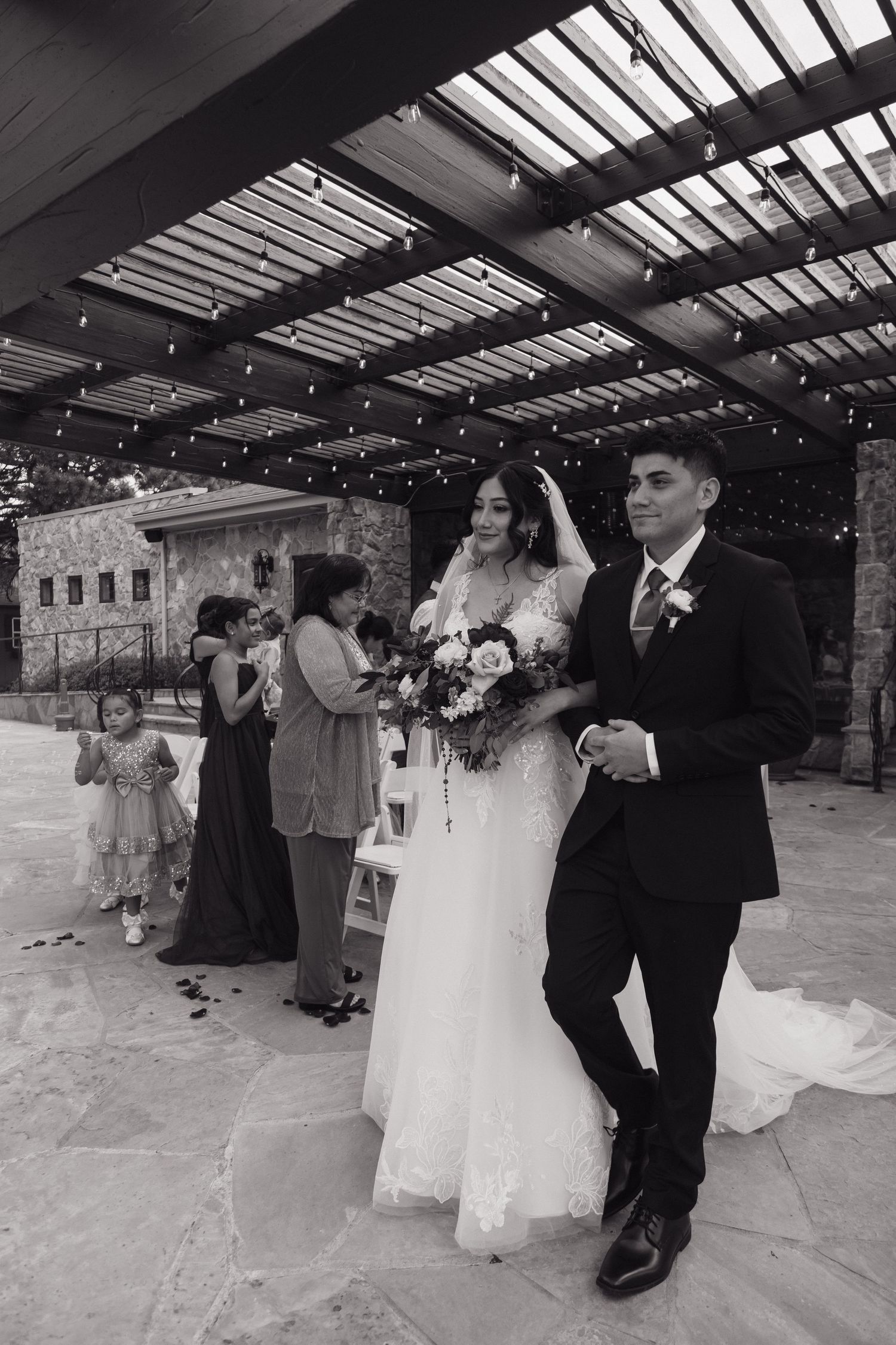 Bride in white lace gown and groom in black suit walk down the aisle after wedding ceremony outdoors.